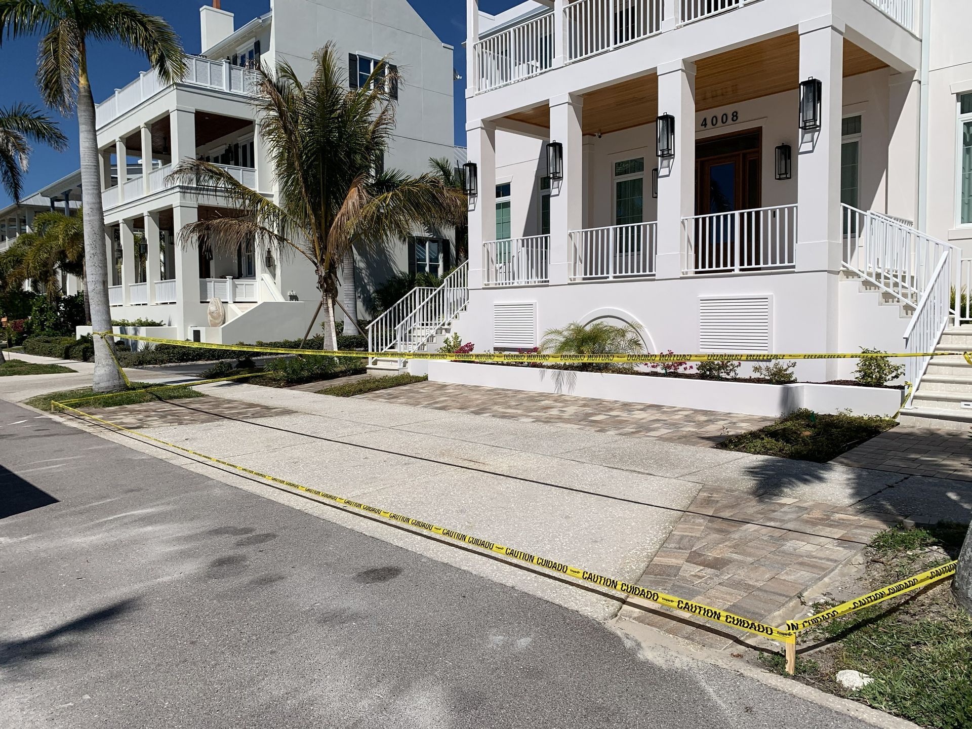 White two-story houses with porches and palm trees along a residential street, viewed from the sidewalk
