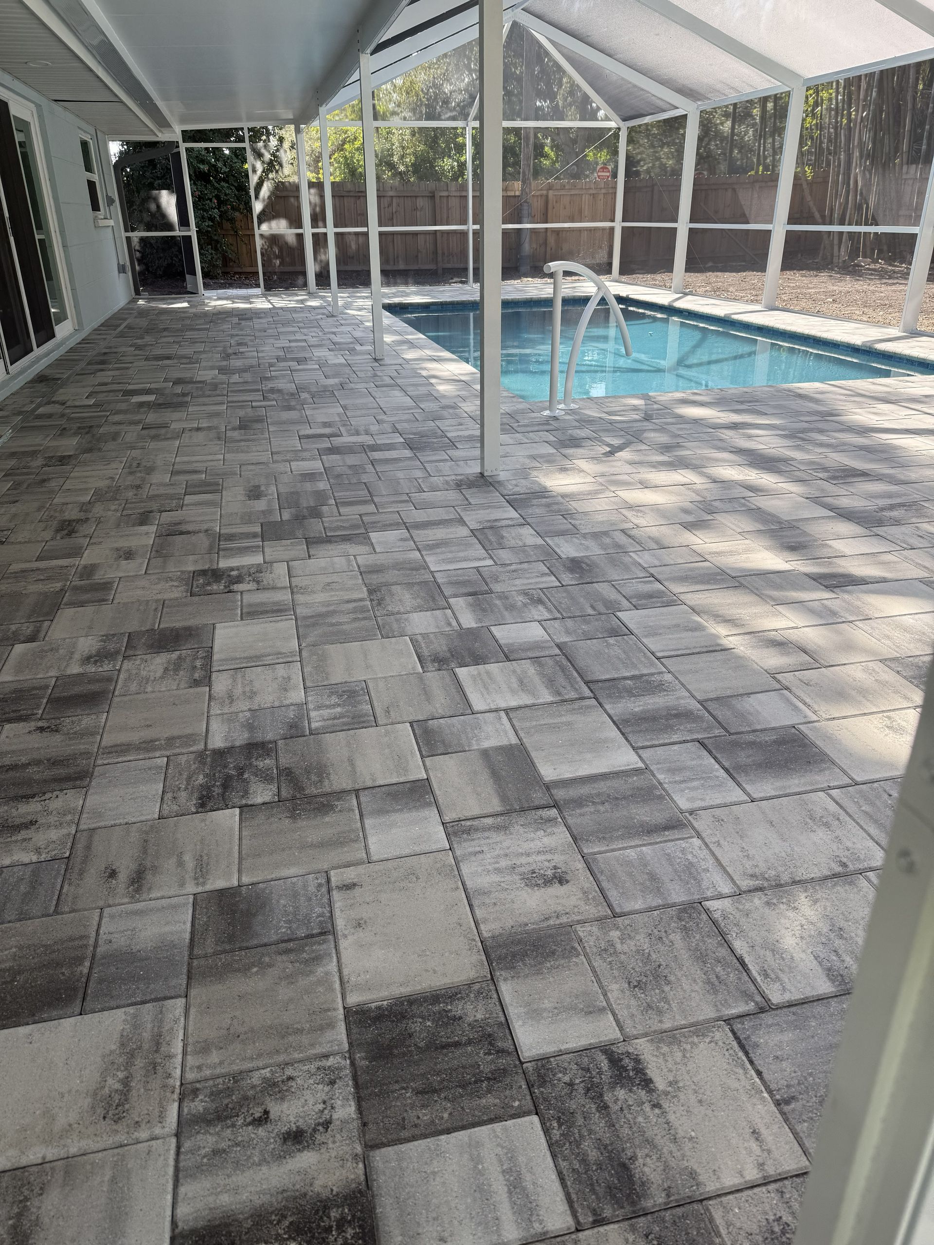 Covered patio with gray pavers beside a screened swimming pool and fenced yard.