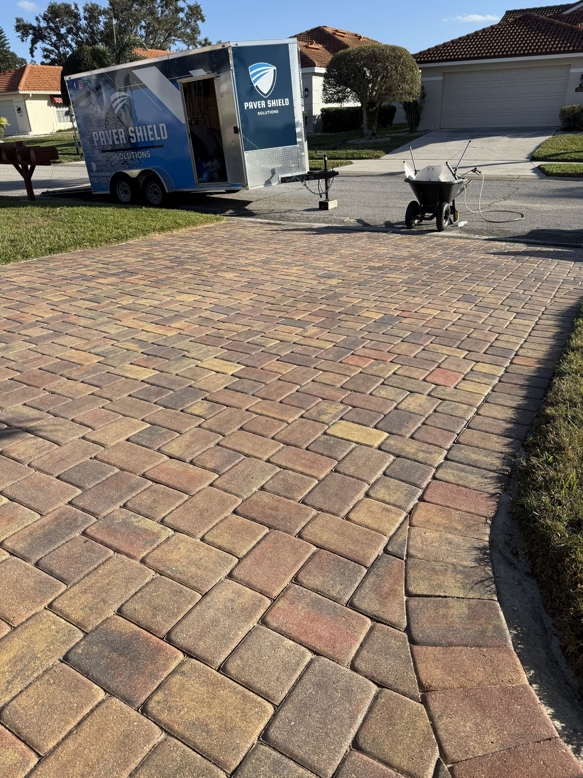 Brick driveway with a blue trailer and a black bin near a house on a sunny day