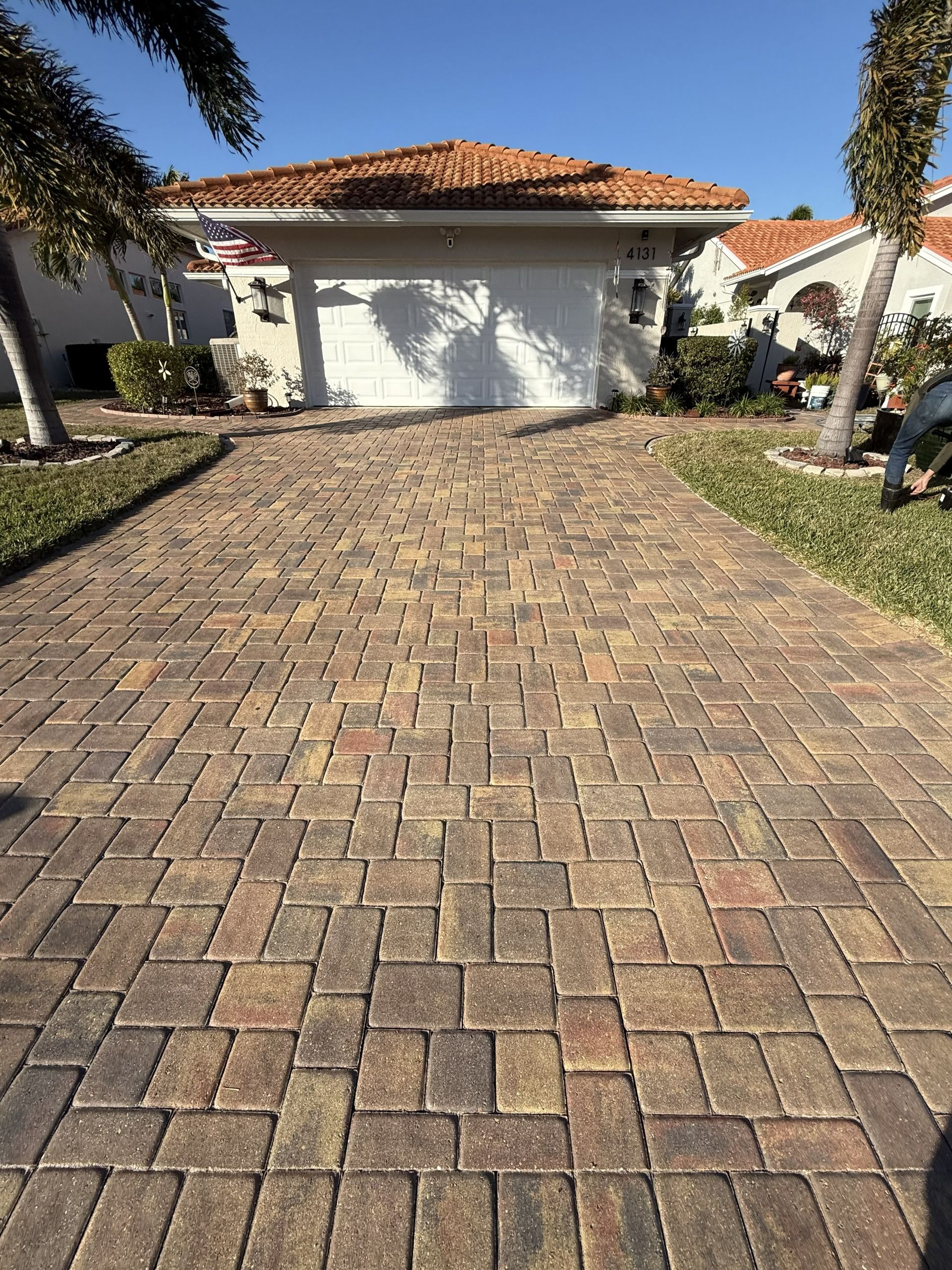 Brick-paved driveway leading to a white garage with a red tile roof and palm trees on either side