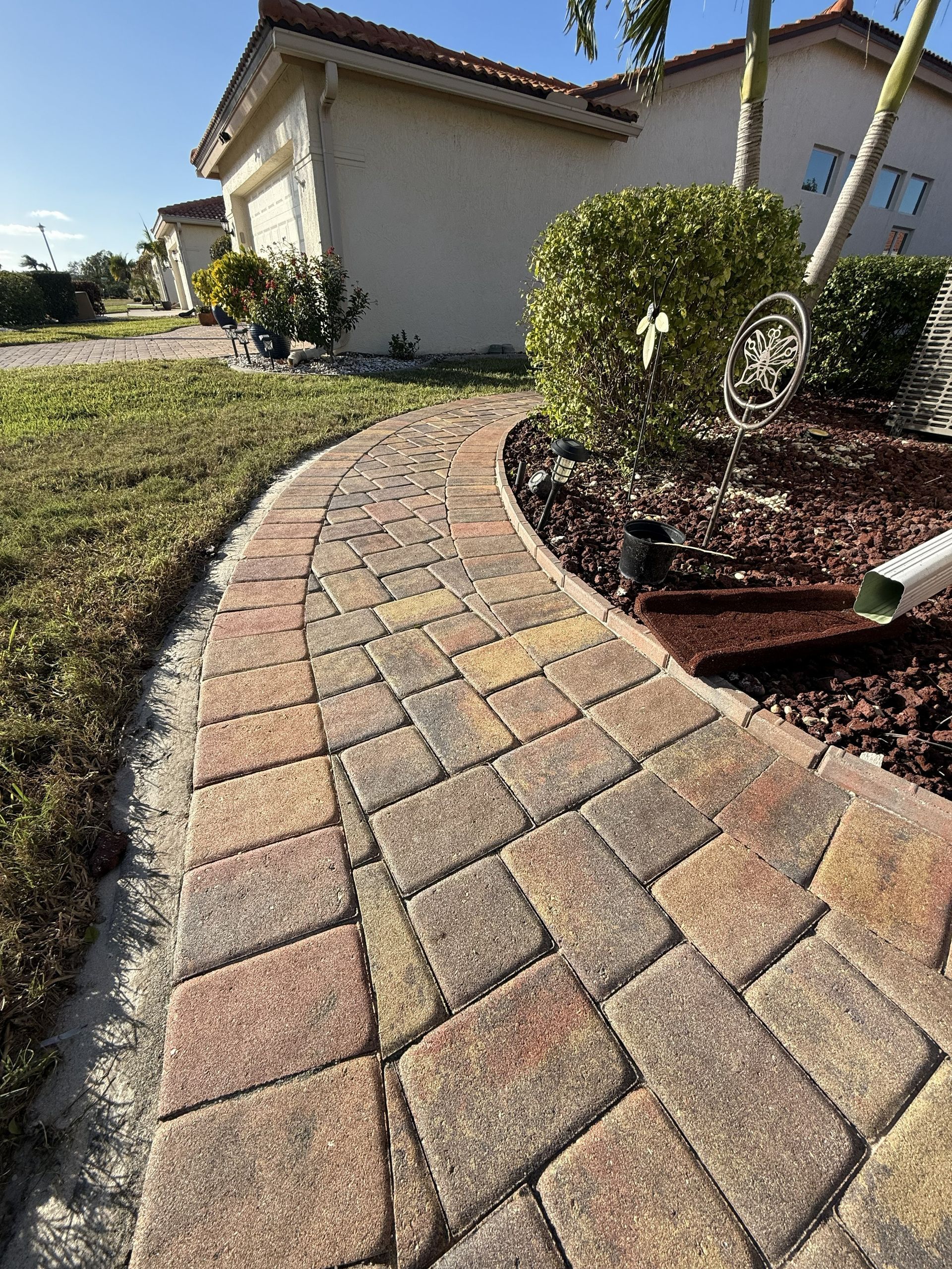 Curved brick paver walkway beside a house and landscaped shrubs in bright sunlight