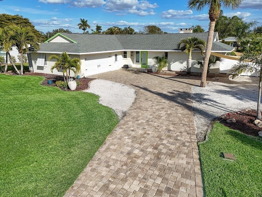 Single-story white house with a curved brick driveway, green lawn, and palm trees under a blue sky