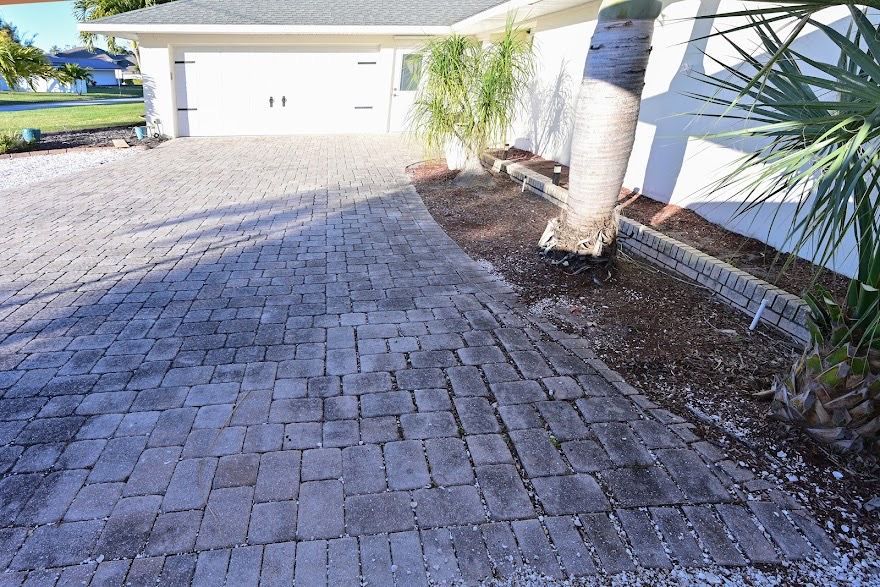 Brick-paved driveway beside a white house and palm trees in bright sunlight