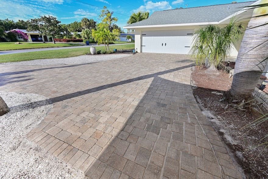 Brick paver driveway leading to a white garage in a sunny suburban home with palm trees and lawn