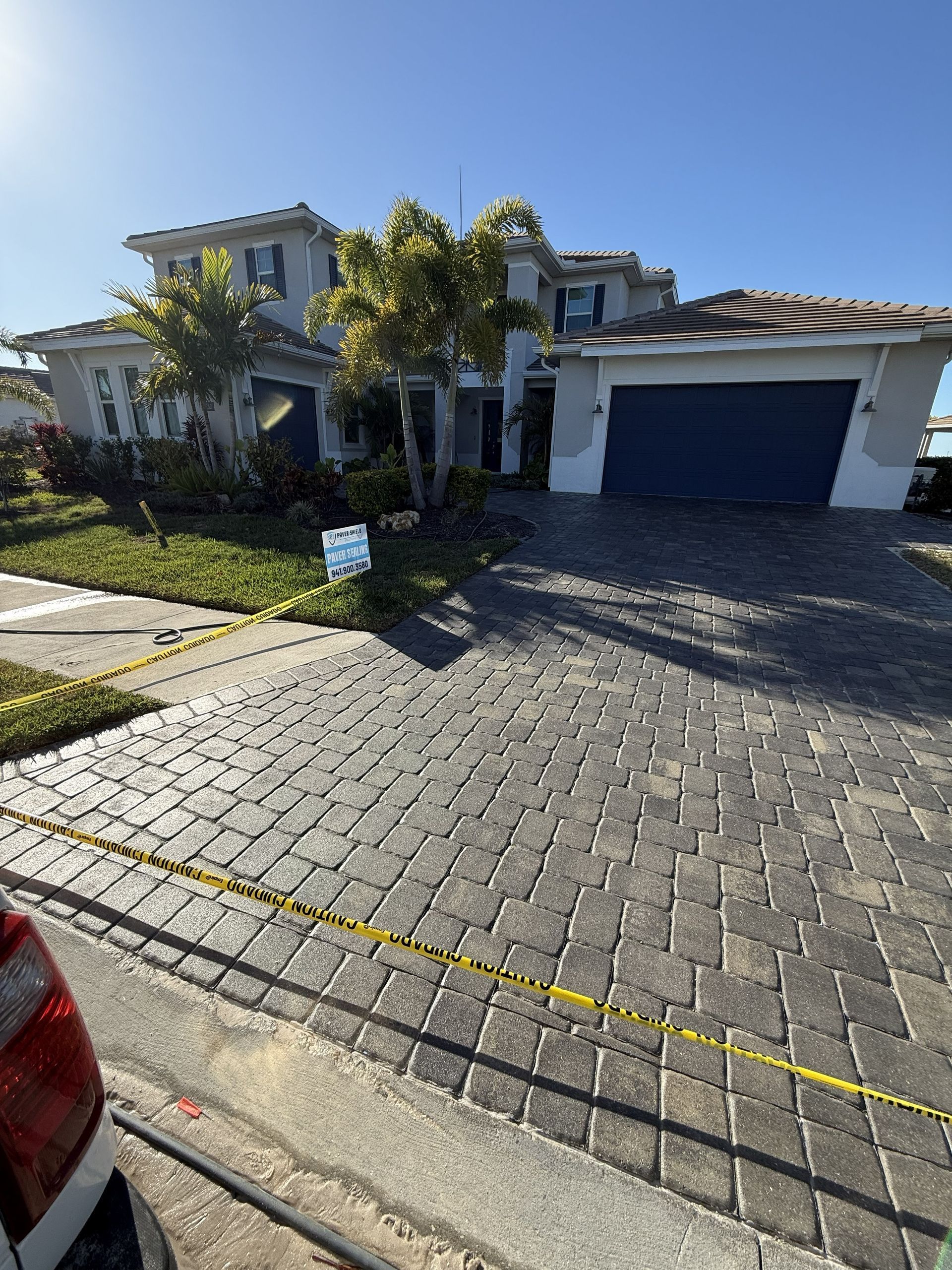 Sunlit suburban house with a blue garage, palm trees, and a brick driveway from the street