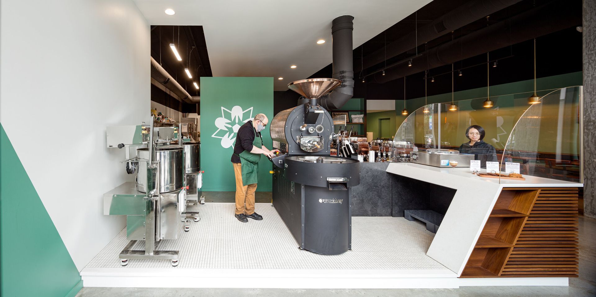 A man is standing in front of a coffee roaster in a coffee shop.