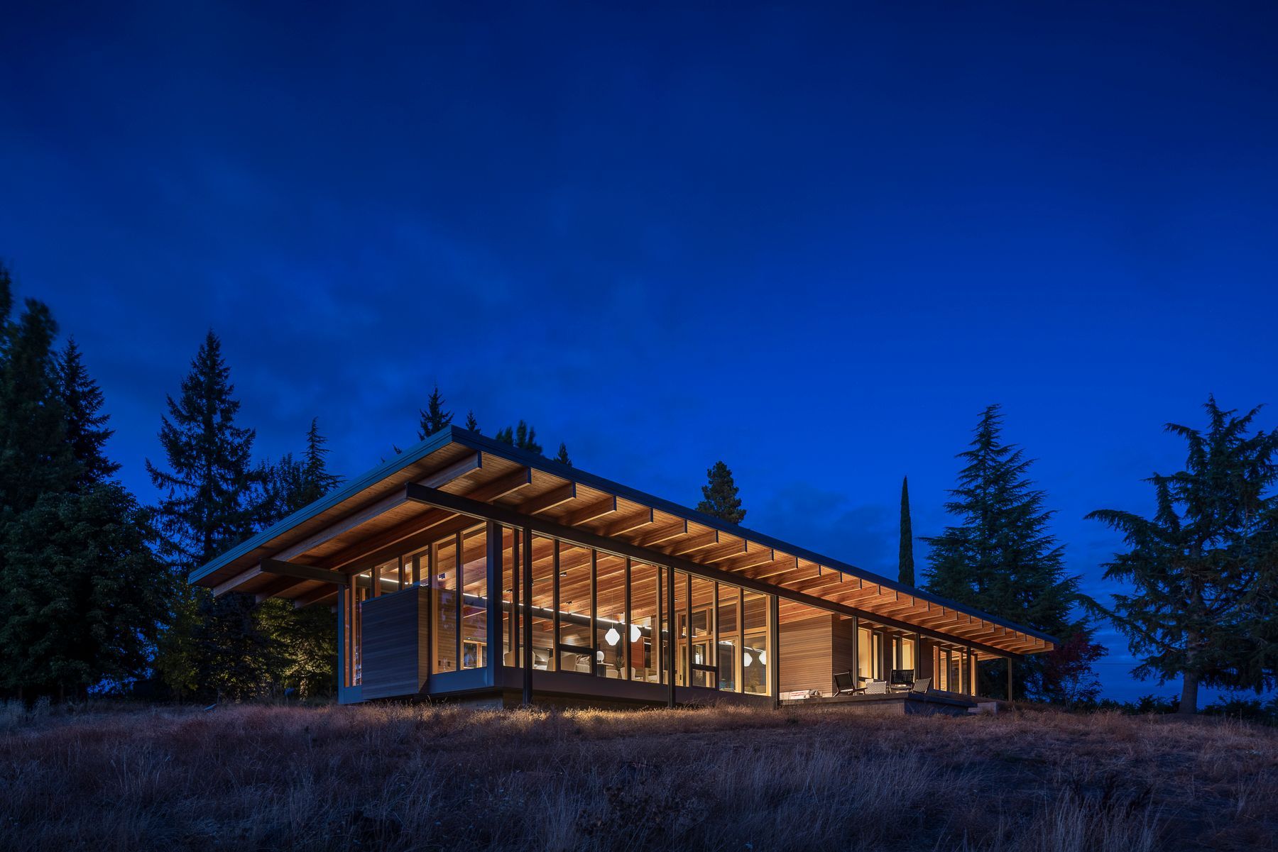A modern wooden house with a lot of windows is lit up at night.