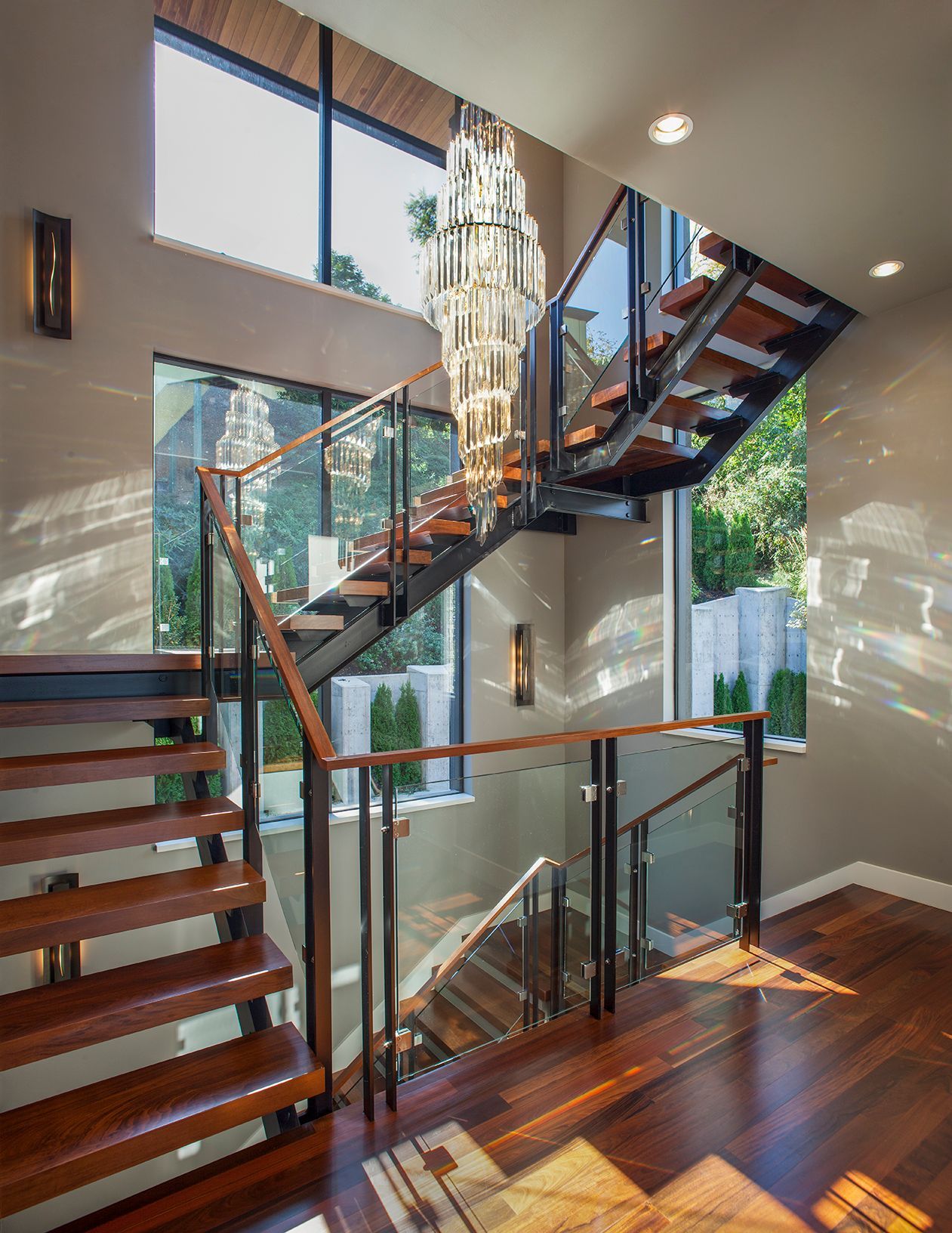 A modern wooden staircase with a glass railing and a chandelier hanging from the ceiling.