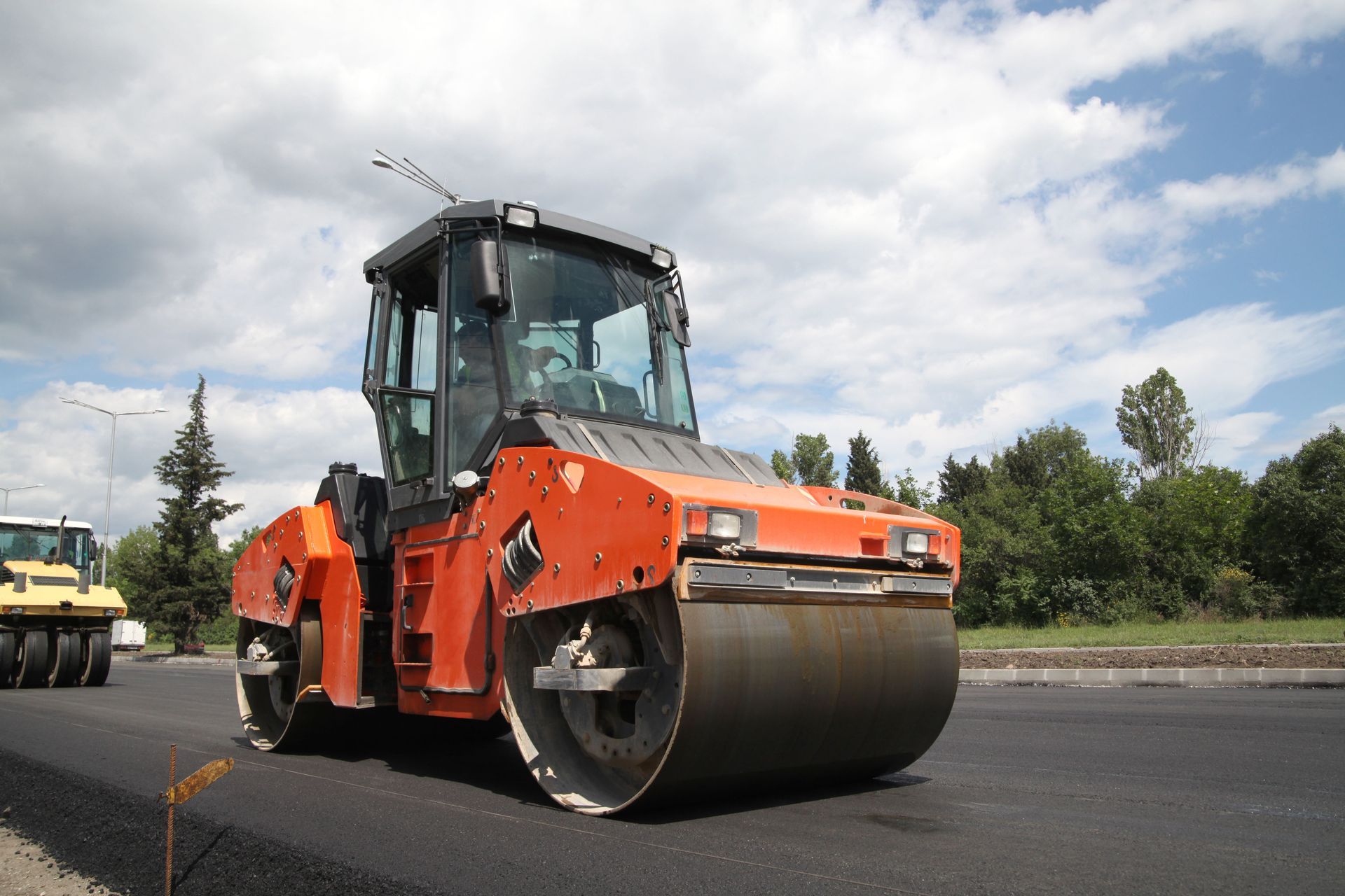 Orange road roller compacting asphalt on a road under a cloudy sky.