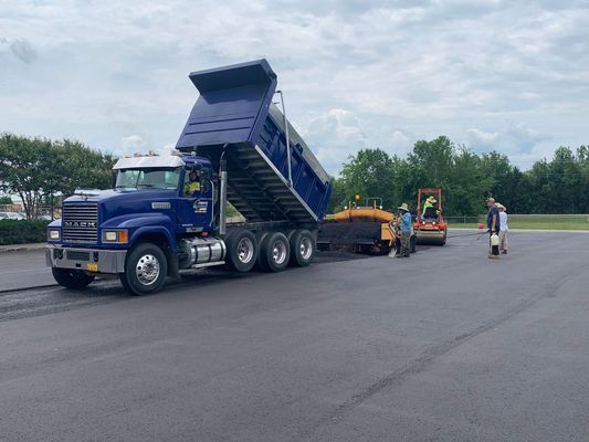 Blue dump truck unloading asphalt onto freshly paved road, with paving machinery and workers visible.