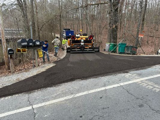 Road crew paving a road with asphalt. Two workers in high-vis vests, machine, mailboxes and trees present.