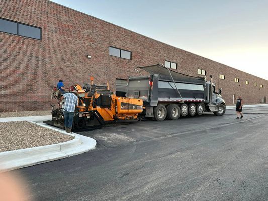 Asphalt paving machine laying pavement next to a brick building. Truck unloading asphalt.