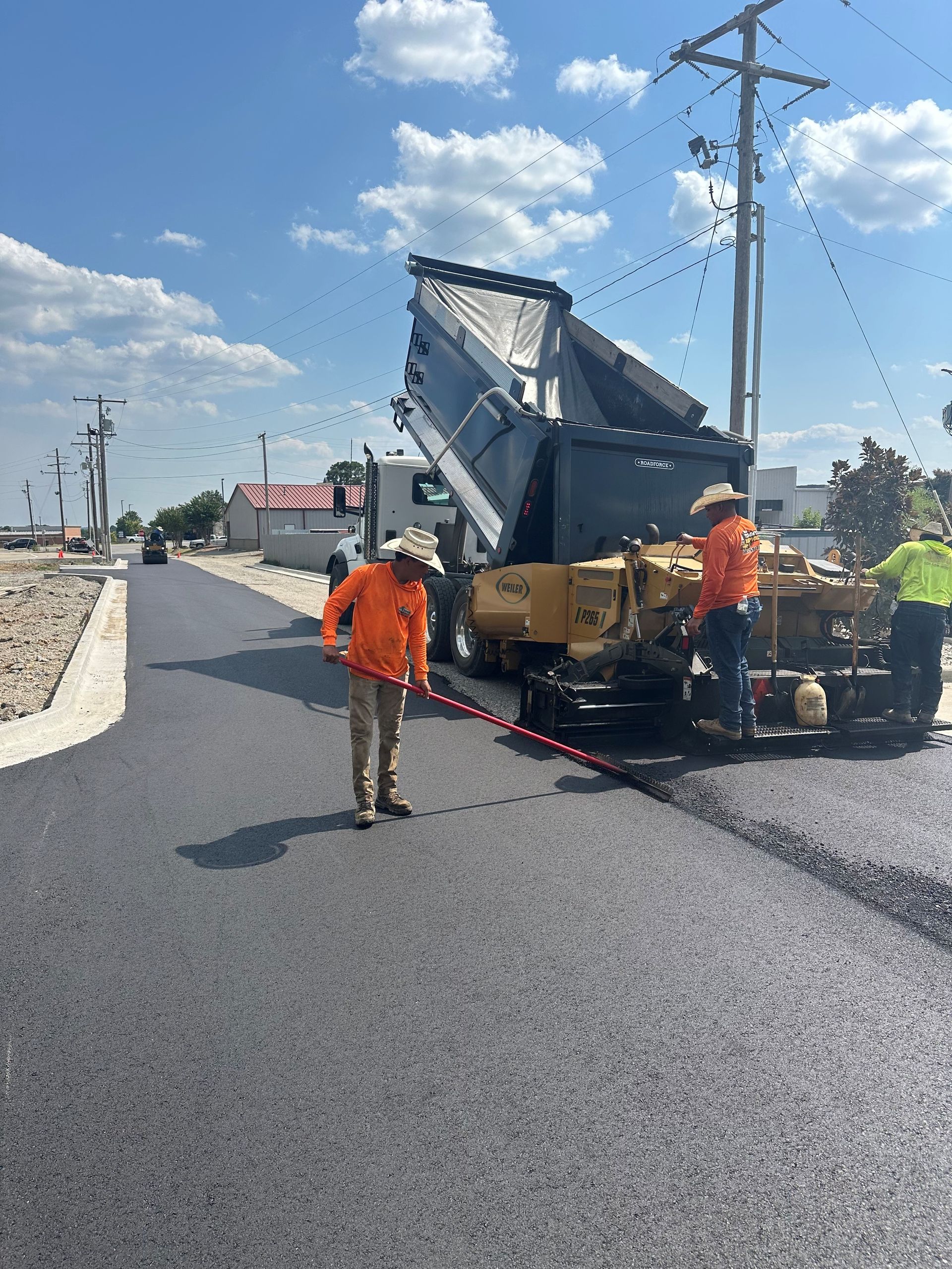 Road crew paving asphalt on a street under a bright sky; dump truck unloading near paving machine.