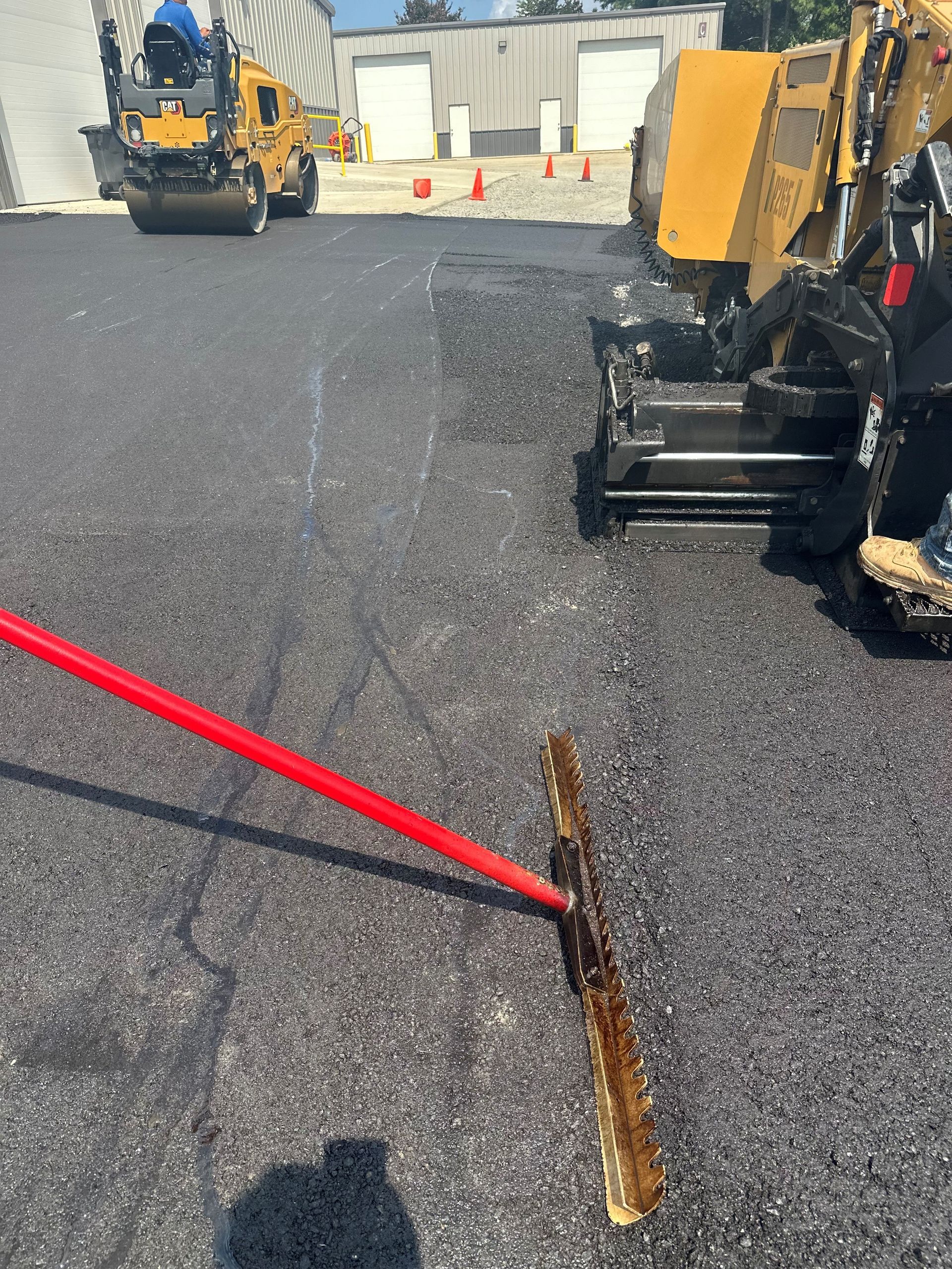Asphalt paving in progress: worker using rake, machinery, roller, and building in background.