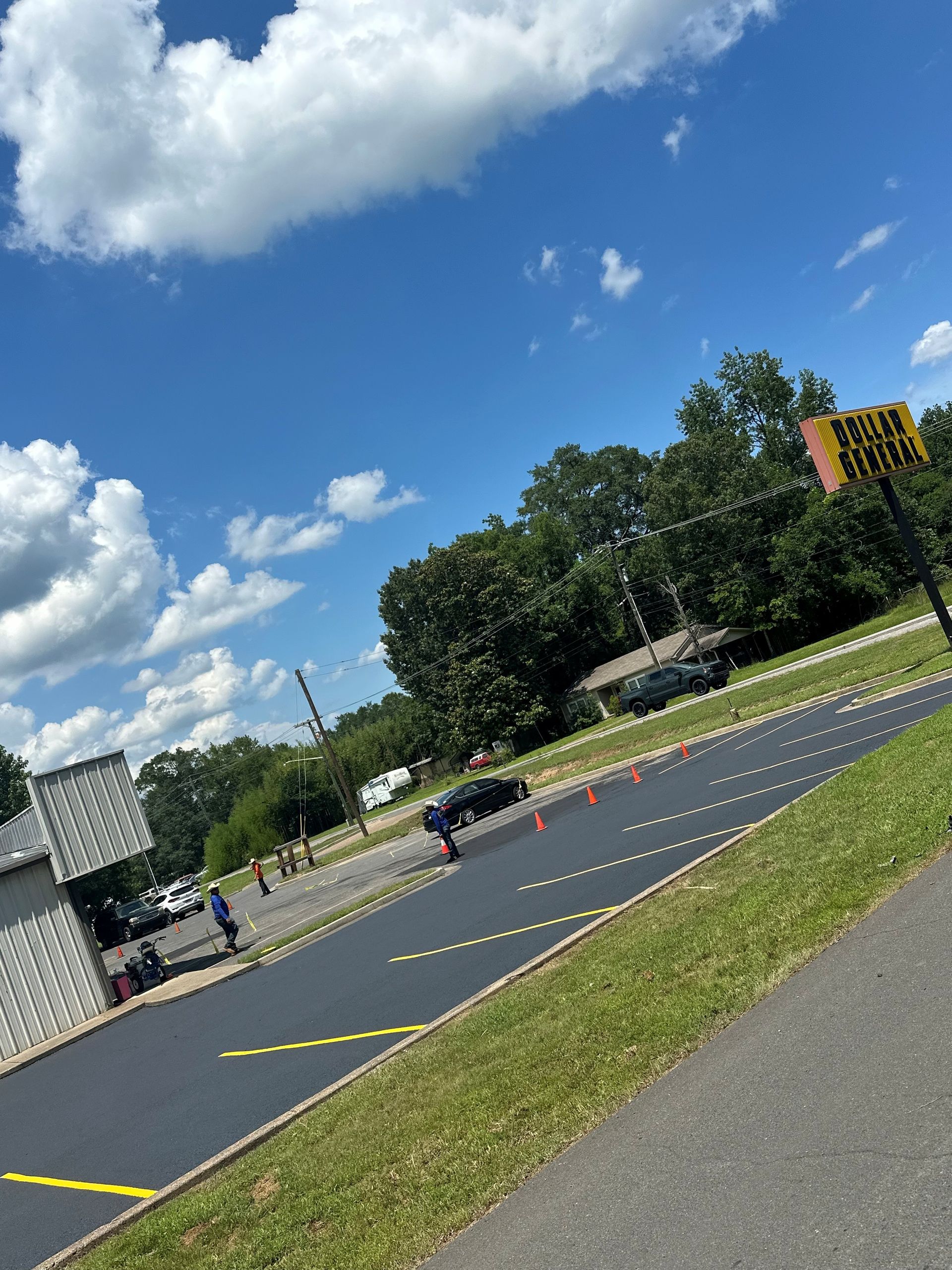 Paved area with cars and orange cones under a bright blue sky; trees and buildings in the background.