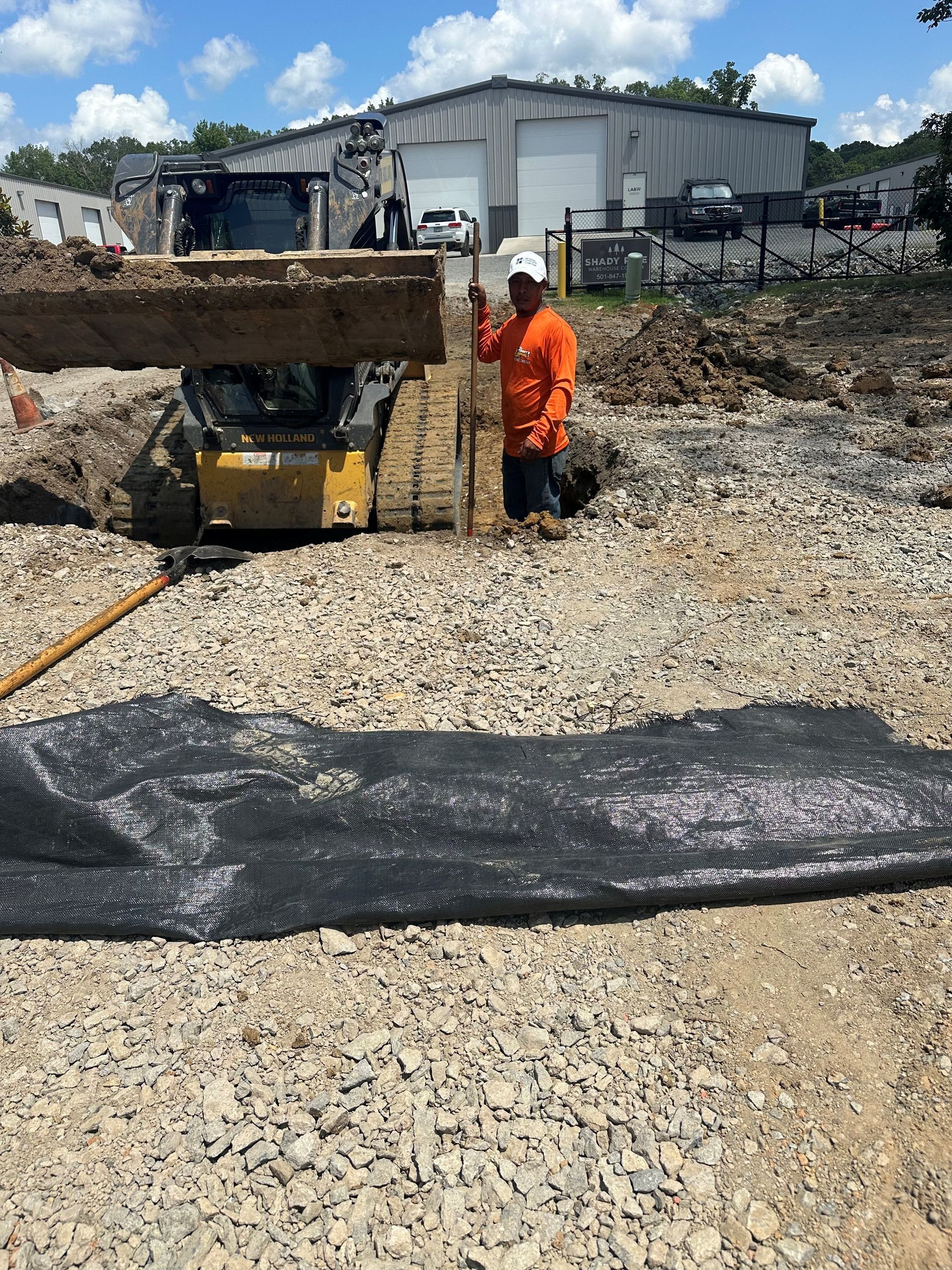 Construction worker in orange shirt kneels near a small digger, holding a plank over a trench. Outdoors.
