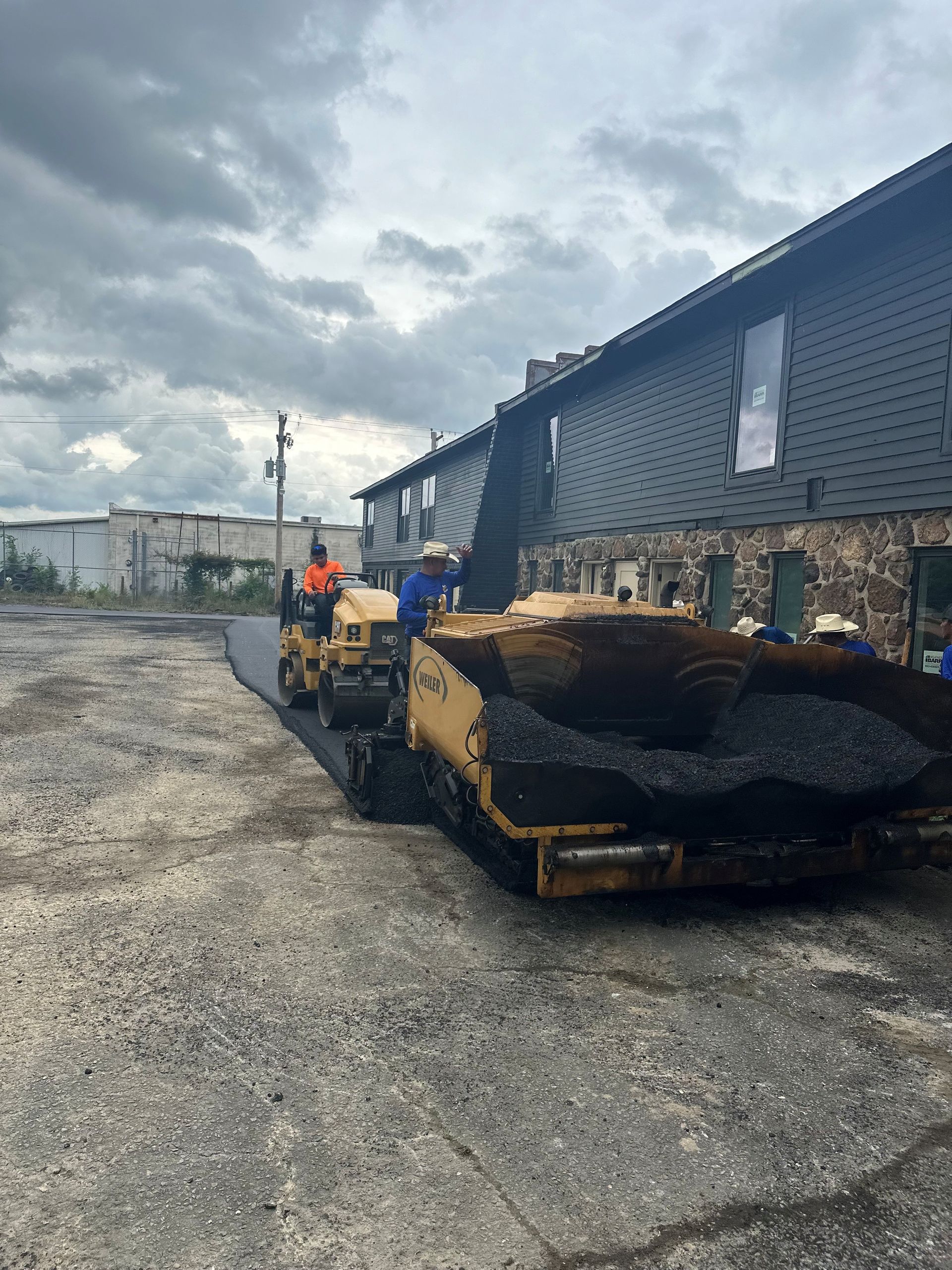 Asphalt paving in progress near a building; crew operating machinery, cloudy sky.