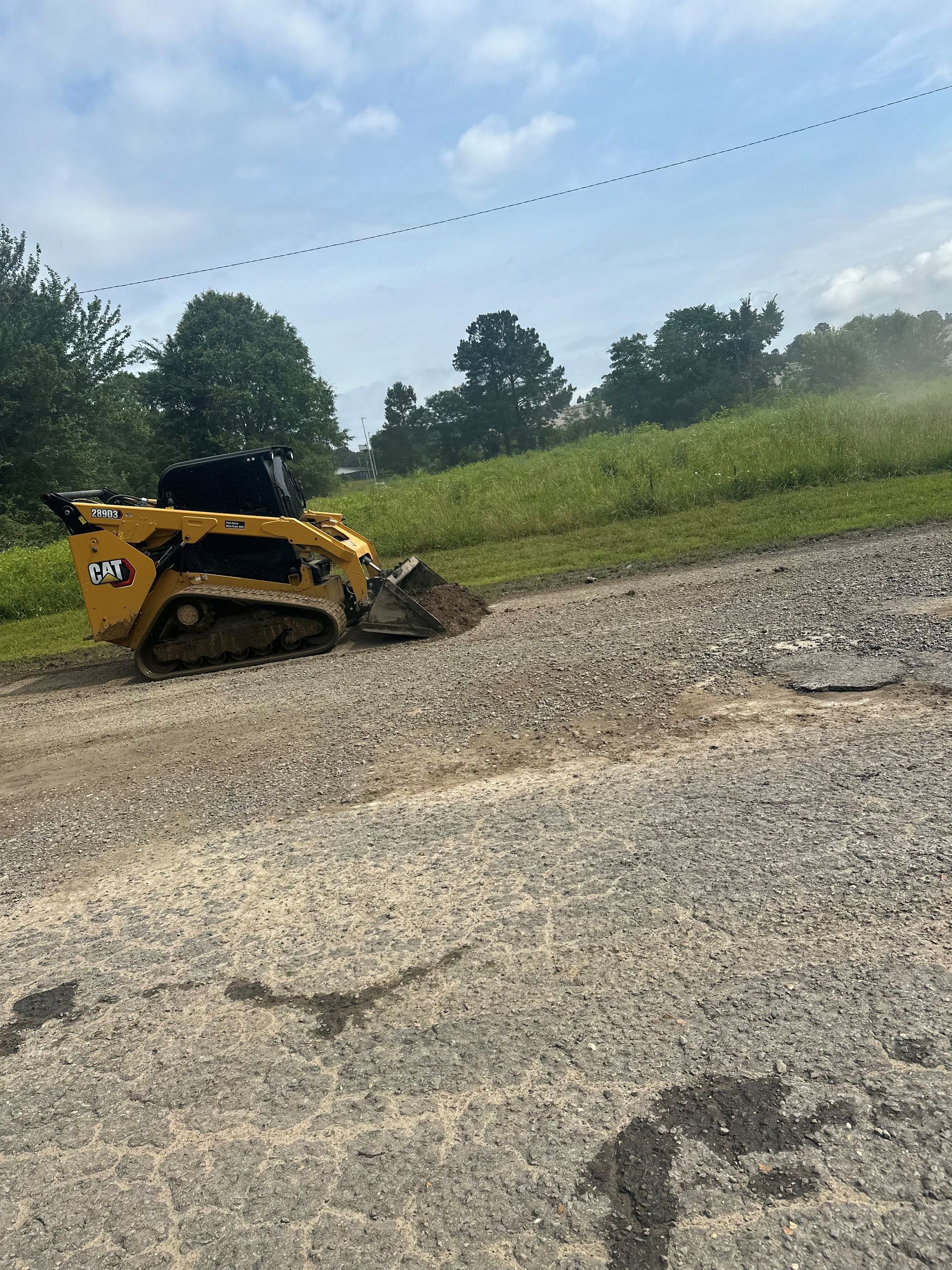 Yellow Caterpillar skid steer on a gravel road with a grassy hill and trees in the background.