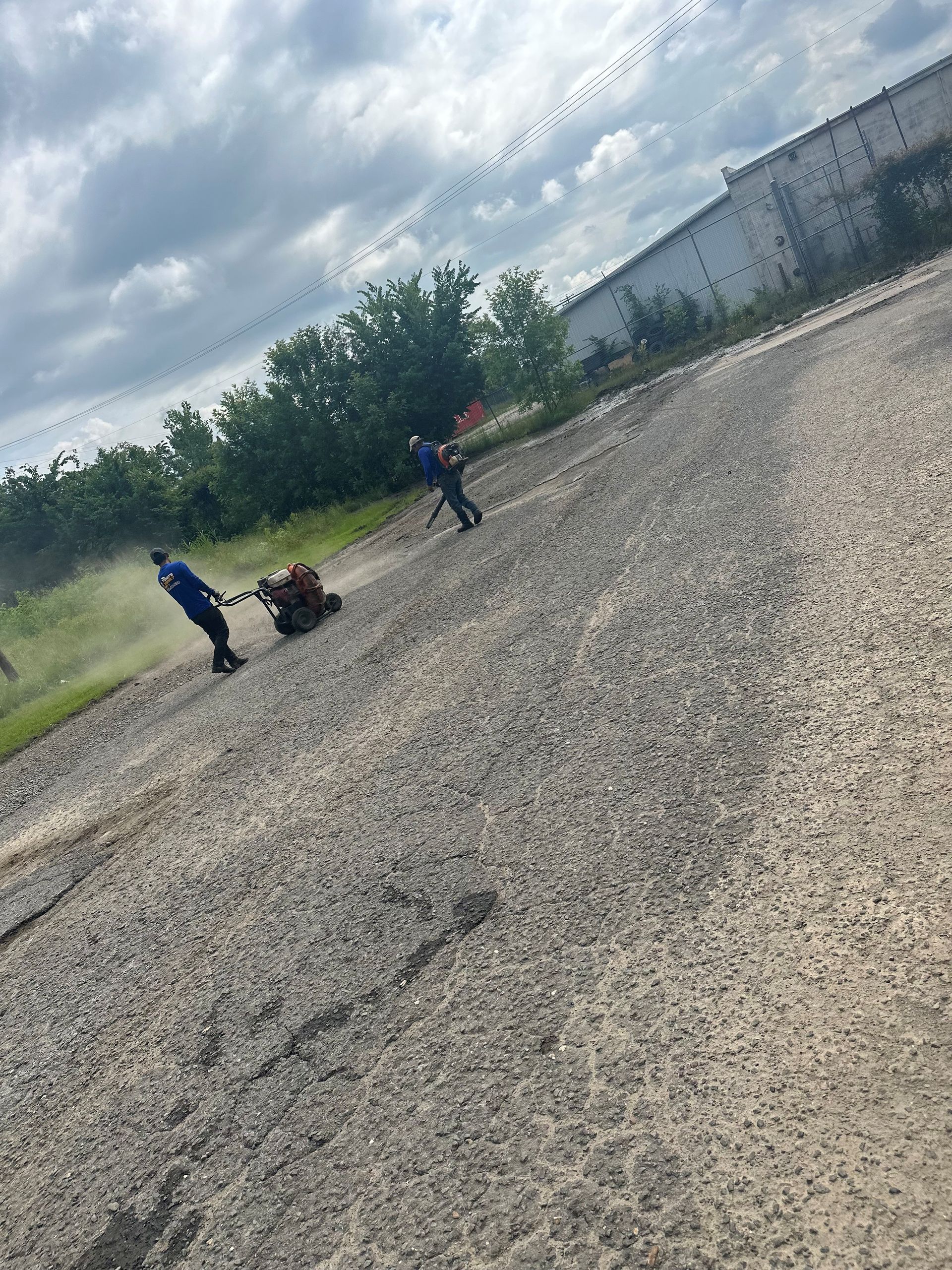 Two people operating a ground machine, possibly for landscaping, on a paved surface. Overcast day.