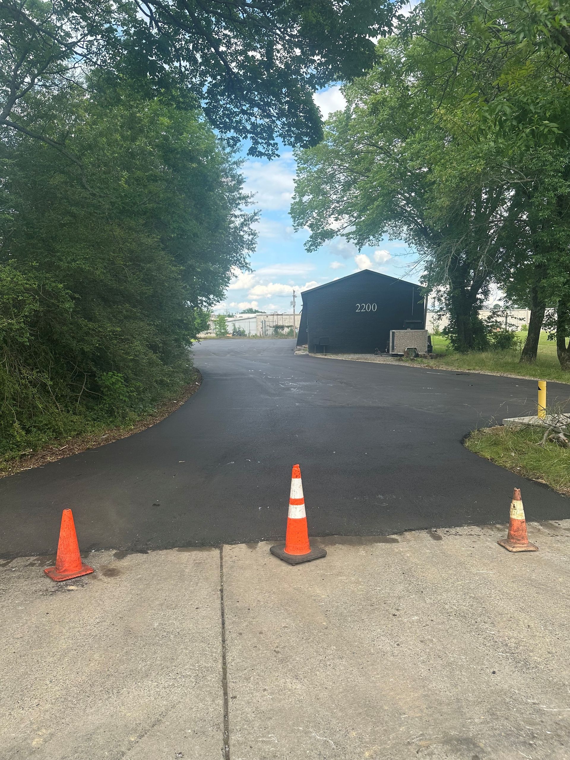 Paved driveway with orange traffic cones and a black building in the distance, flanked by trees on a sunny day.