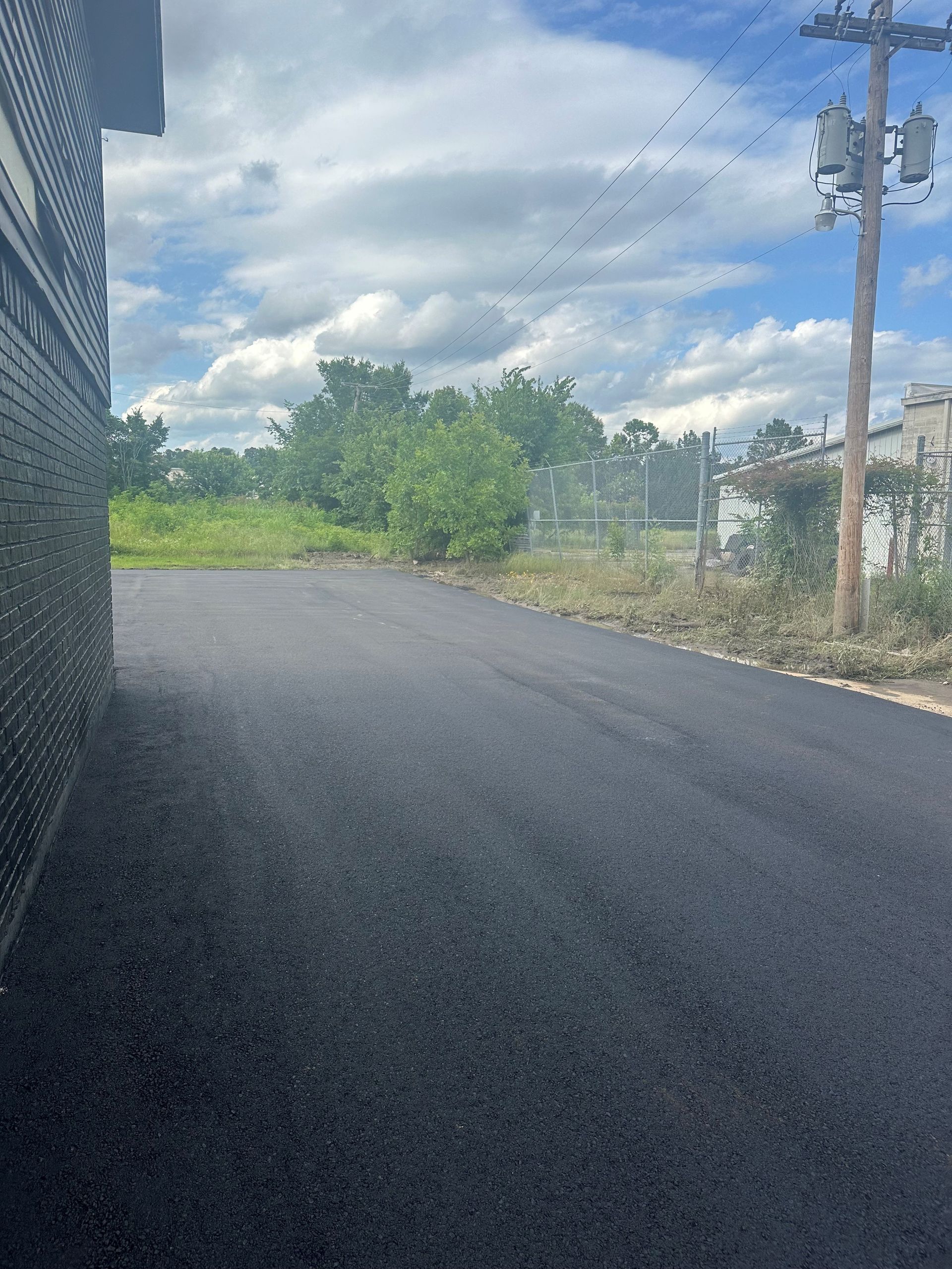 Newly paved asphalt road next to a brick building and green foliage under a cloudy sky.