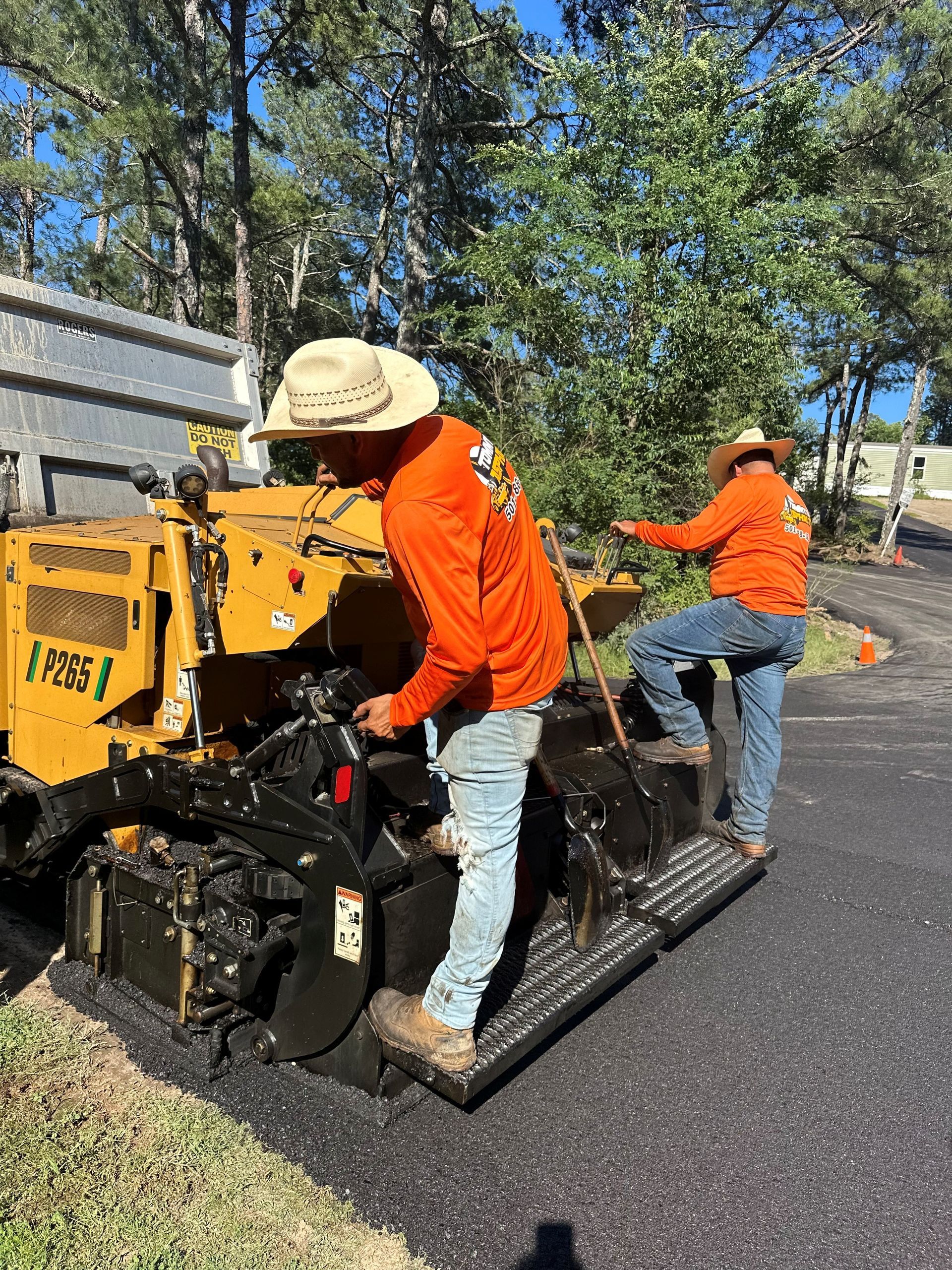 Two workers paving road with yellow asphalt machine, wearing orange shirts, and cowboy hats.