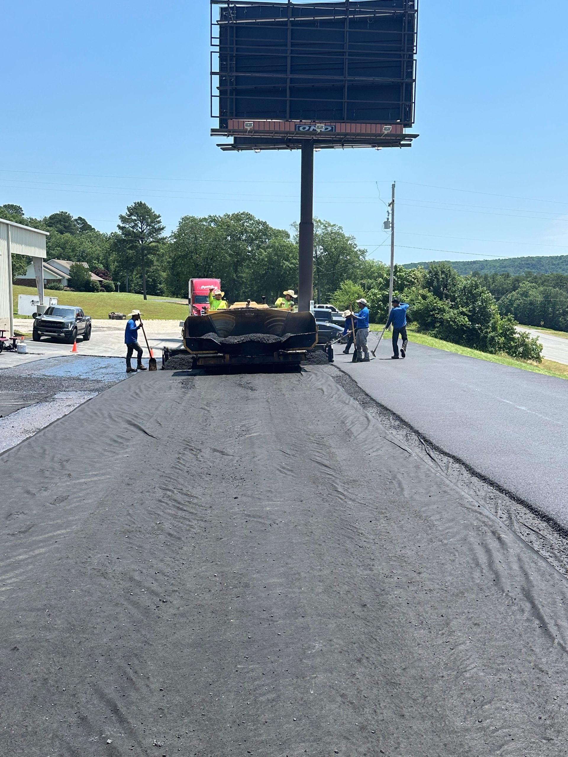 Road paving in progress; workers near billboard, asphalt on roadway, blue sky.