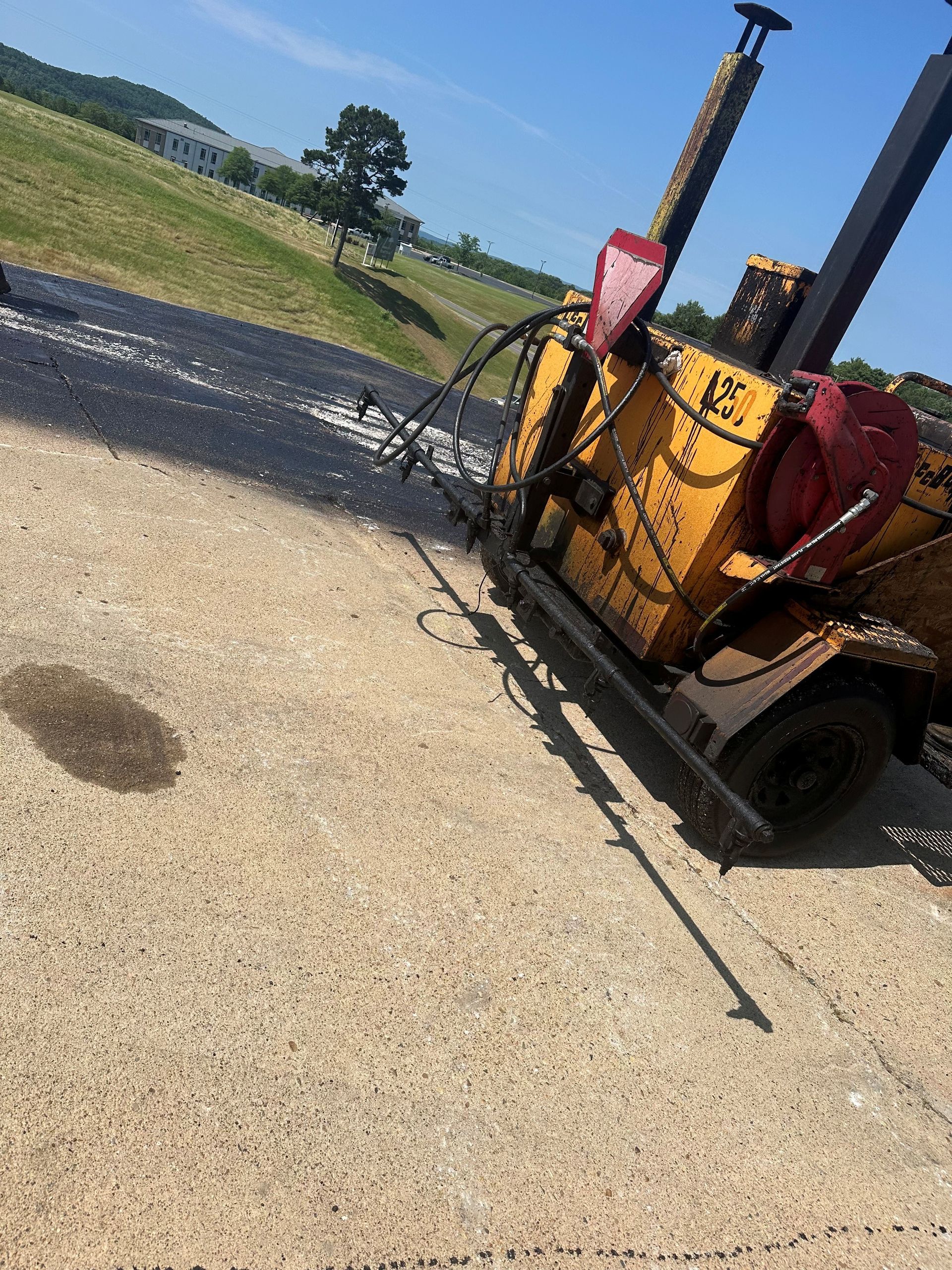 Yellow road repair machine on pavement, casting shadow. Green grass and blue sky background.
