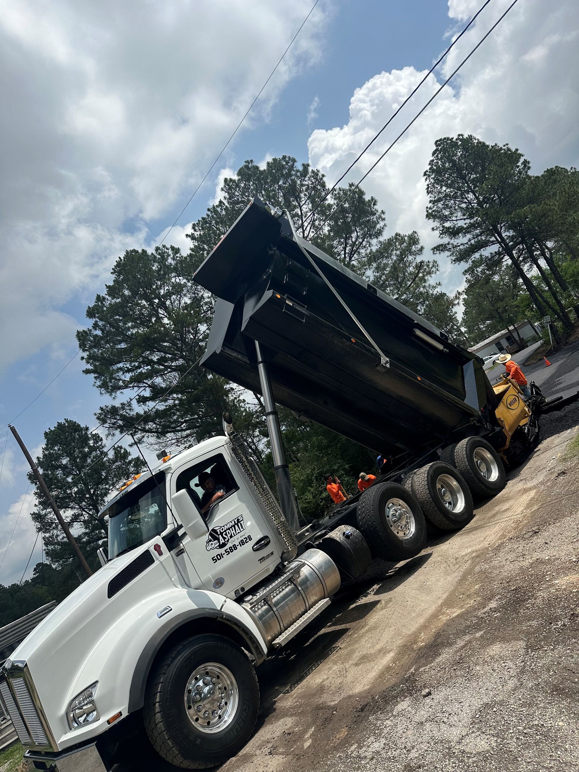 White dump truck with its bed raised, asphalt dumping against a backdrop of trees and sky.