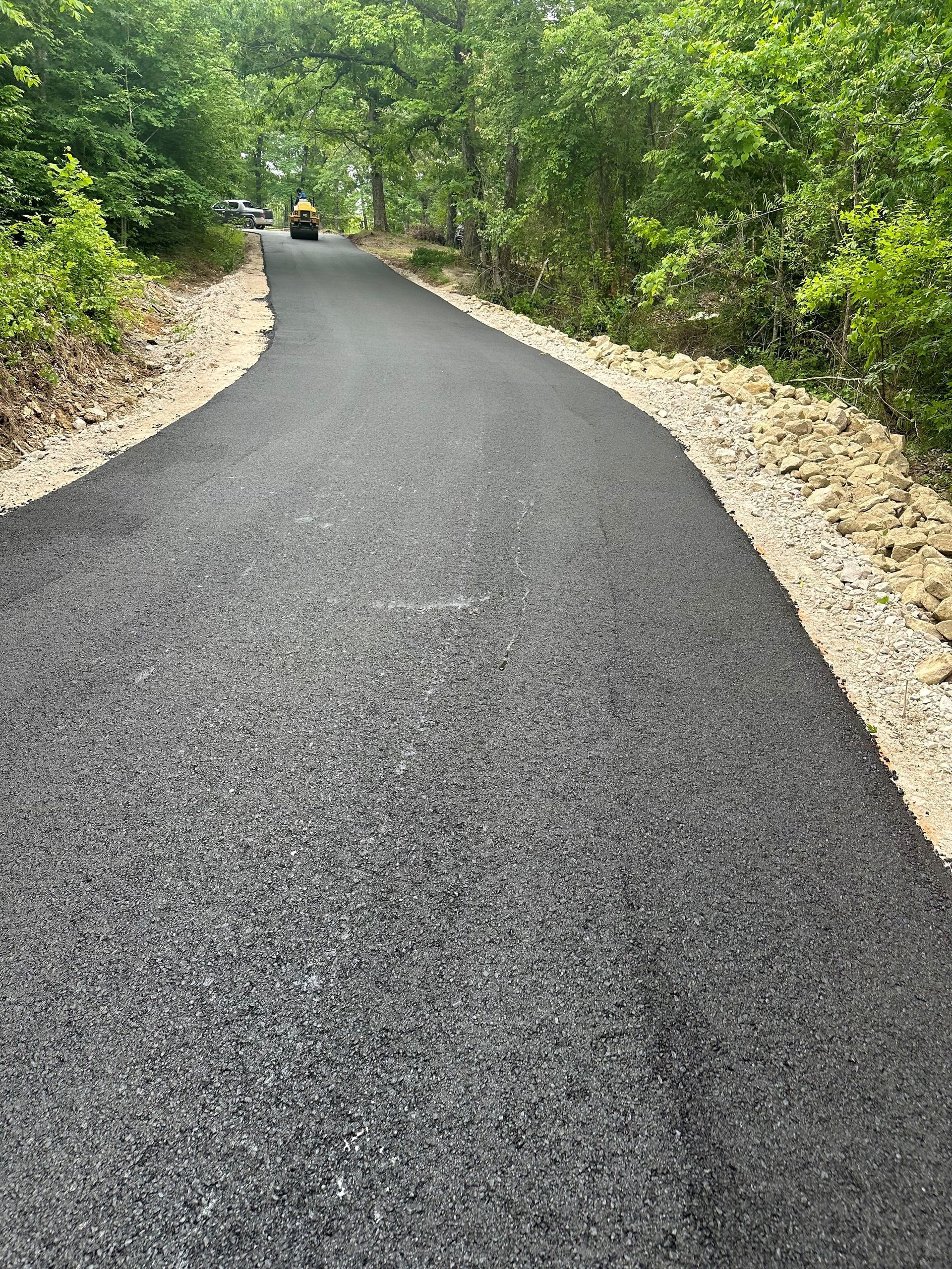 Paved asphalt driveway curves through a wooded area, bordered by gravel and trees.