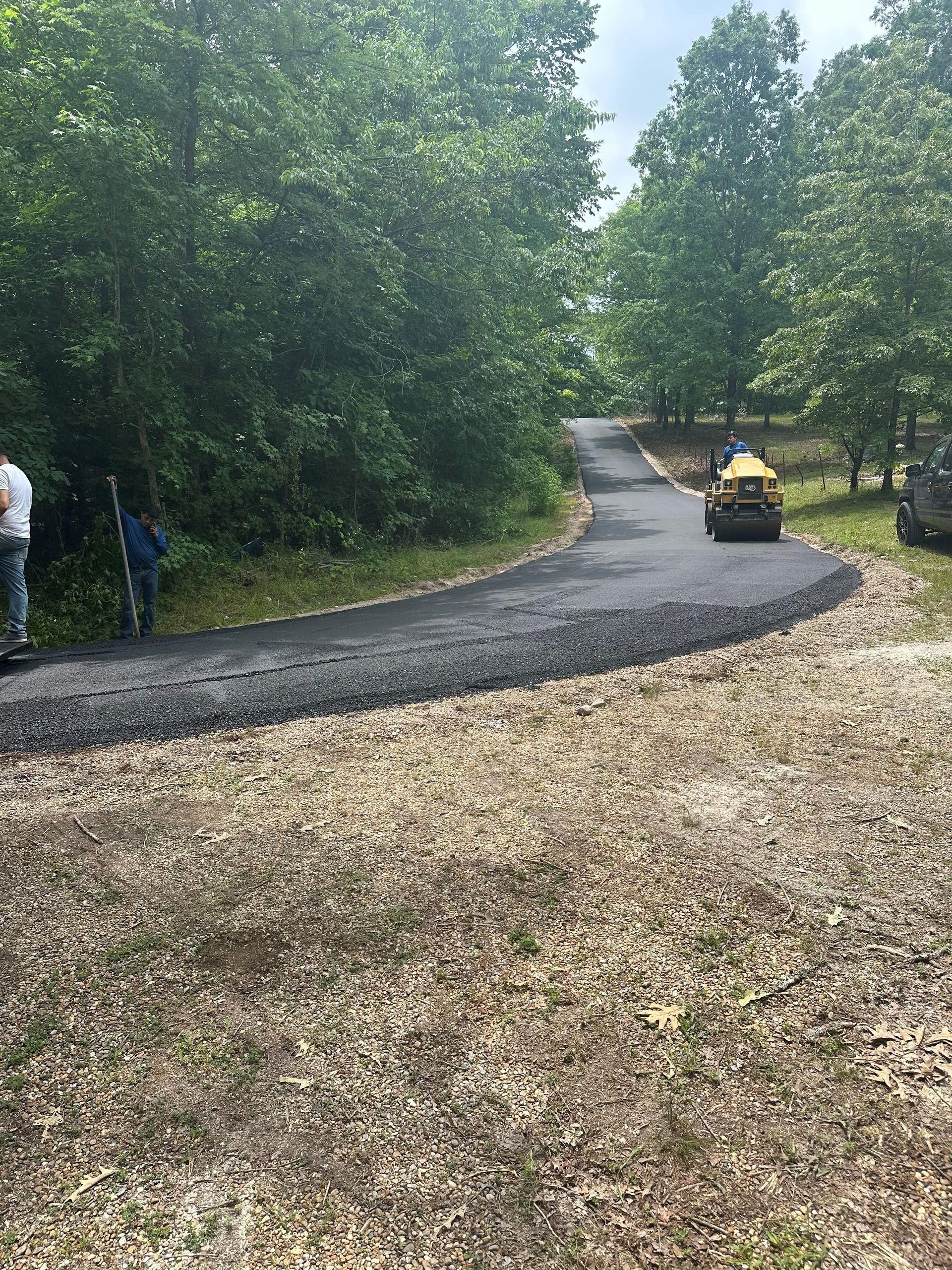 Asphalt road being paved with a roller machine in a wooded area; workers are present.