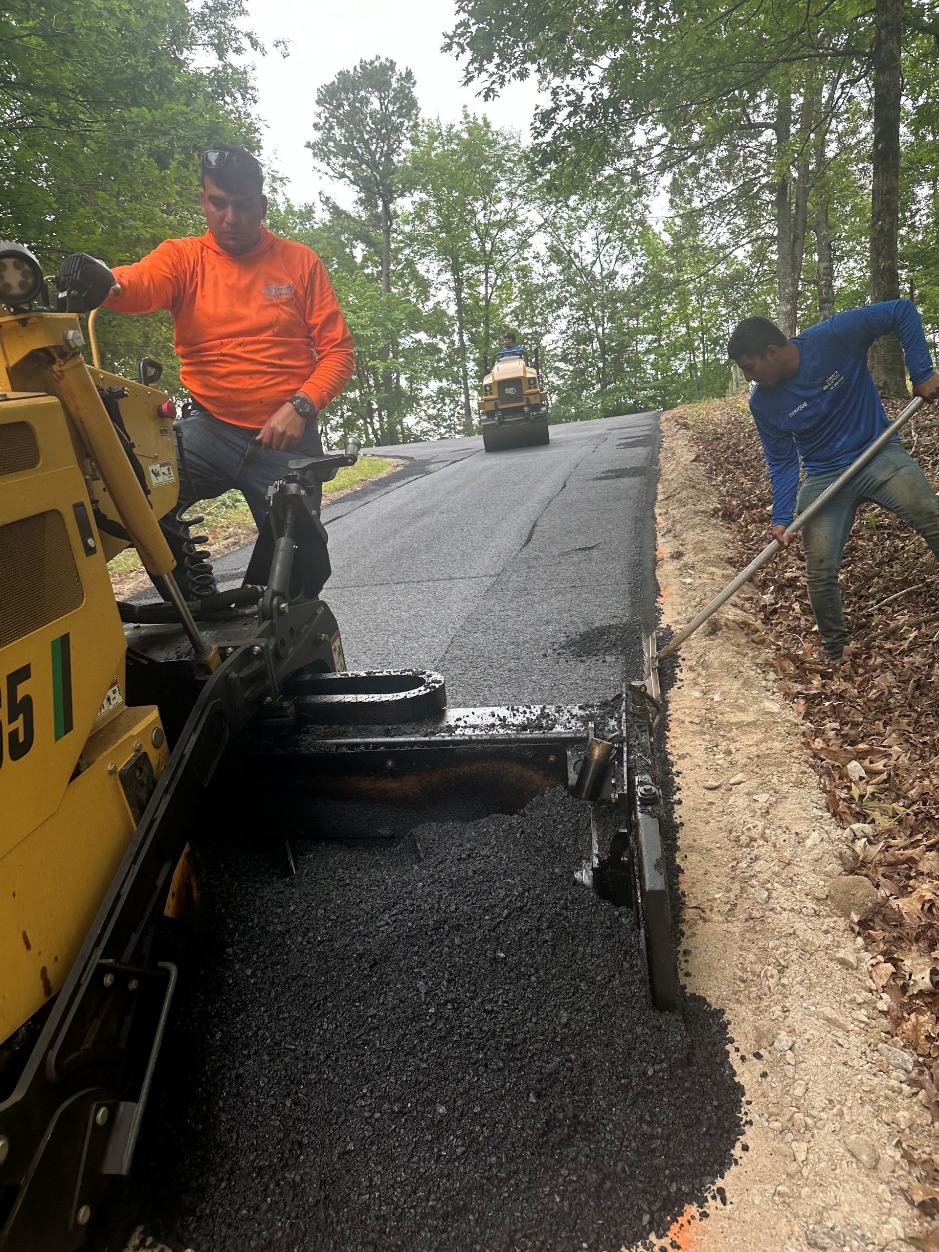 Workers paving a road; one operates machinery, another rakes asphalt. Roadside trees in the background.