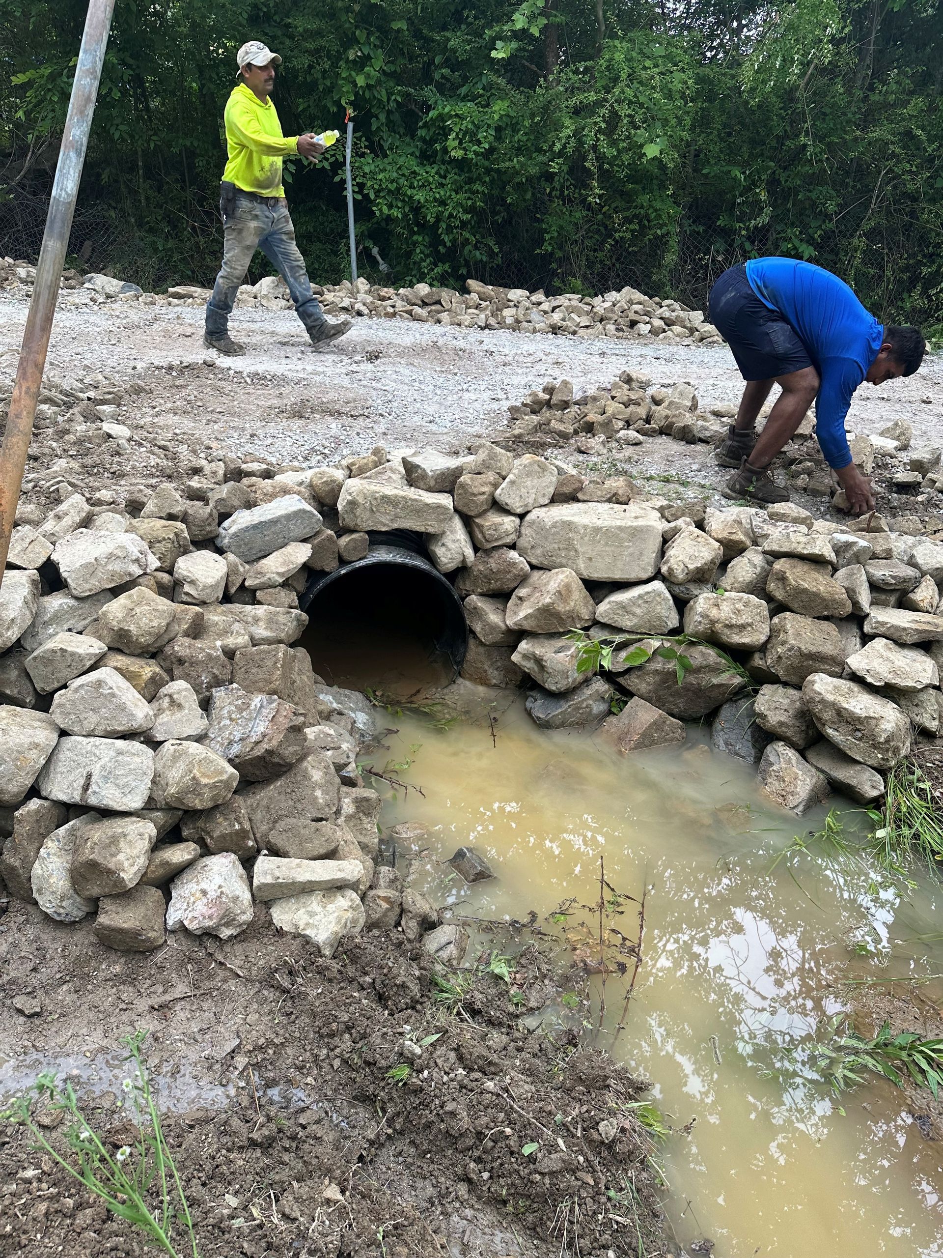 Workers building a stone culvert around a pipe. One man walks; another bends over the construction. Muddy water flows.
