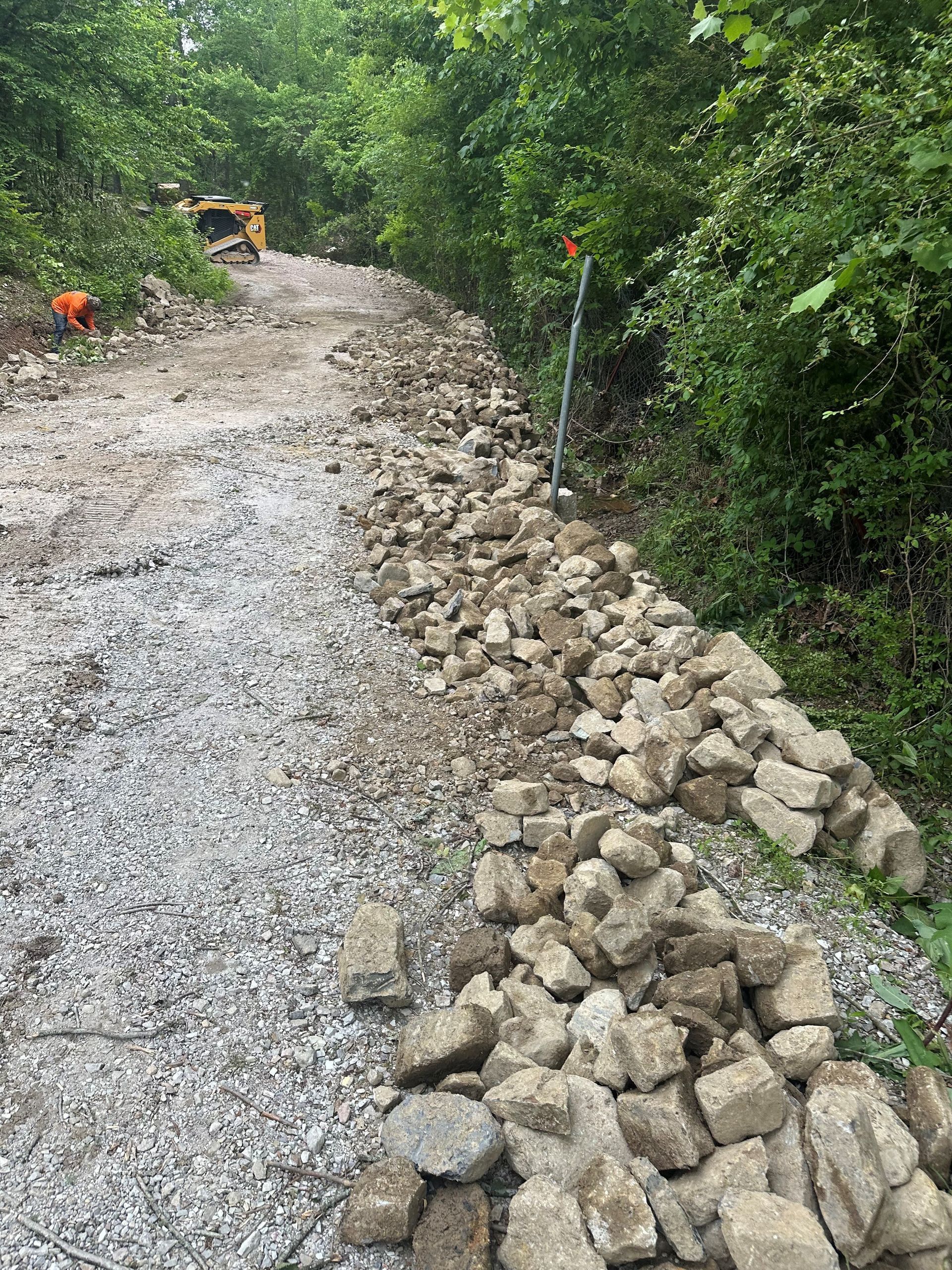 Gravel road under construction. Large rocks line the edge. Yellow construction vehicle in the distance.