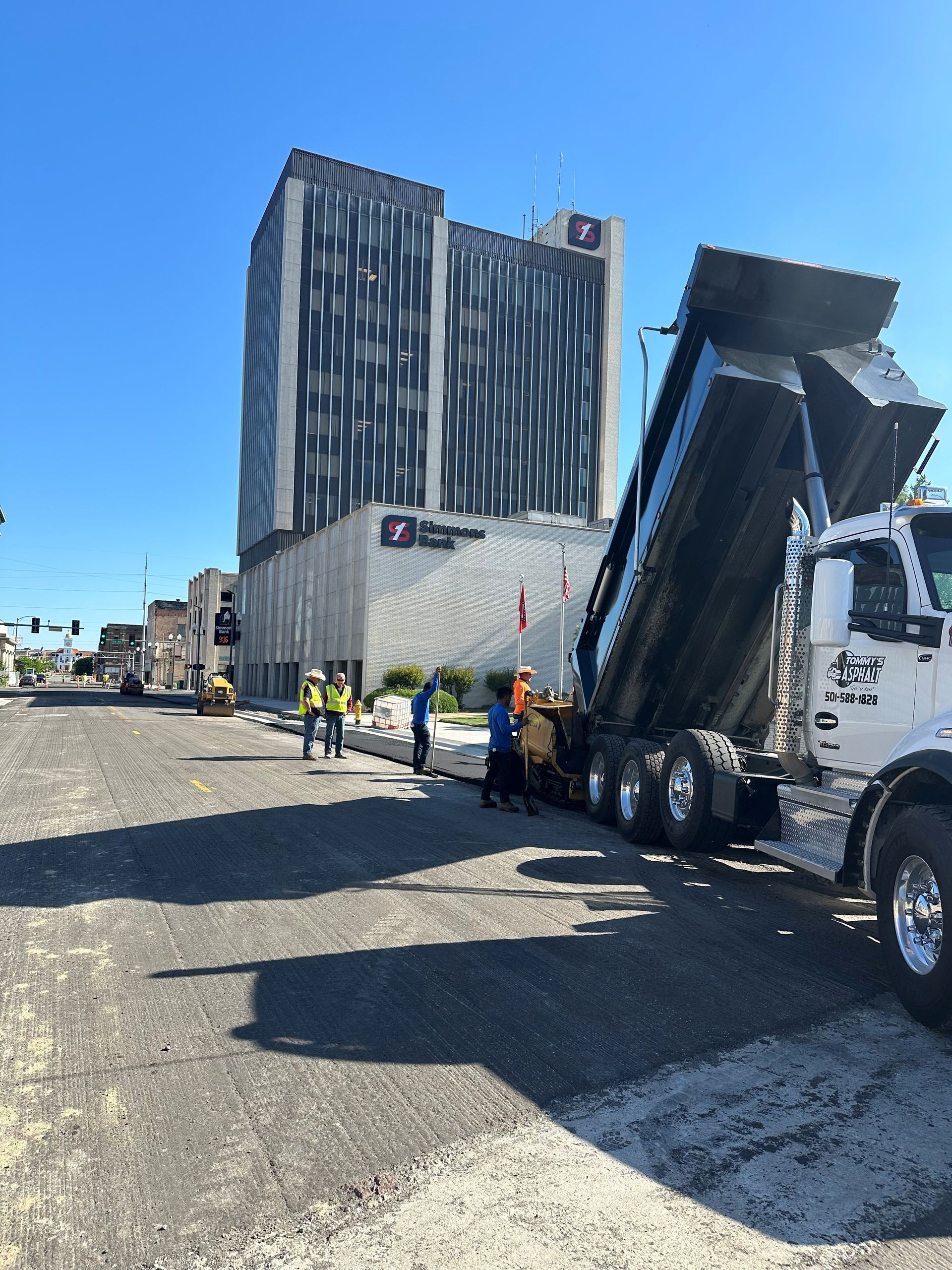 Pavement construction; dump truck unloading asphalt on city street in front of tall building. Construction workers oversee.