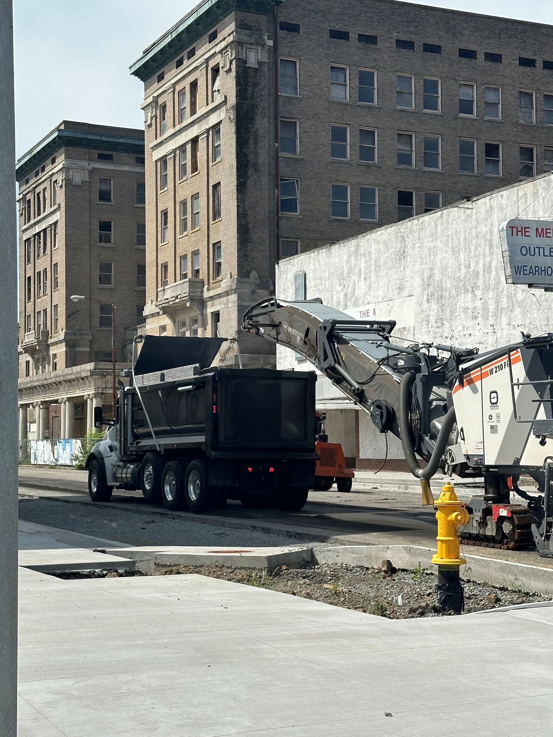 Dump truck loading debris from a road milling machine on a city street, with old brick buildings in the background.