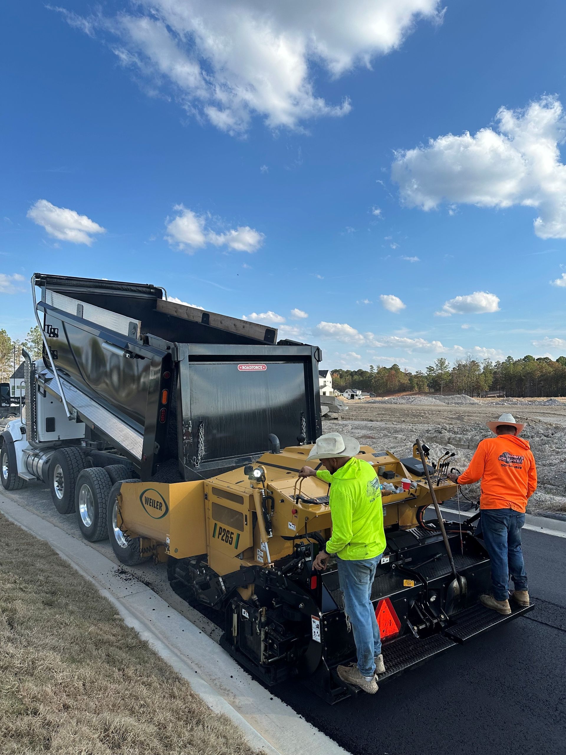 Dump truck pouring asphalt into a paving machine on a road, two workers. Blue sky, sunny day.