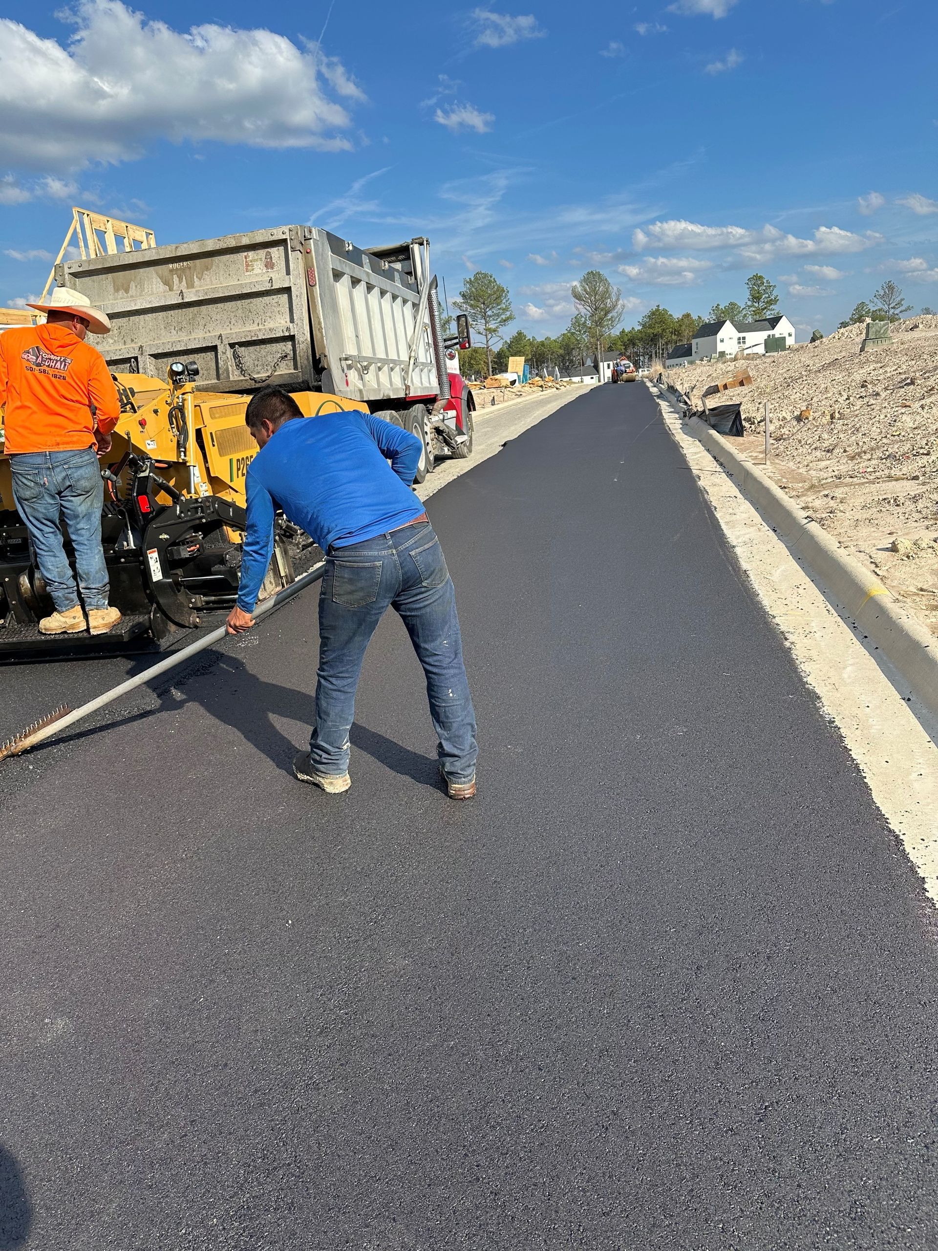 Construction workers paving a road with fresh asphalt. One worker uses a tool on the new surface. Bright blue sky.