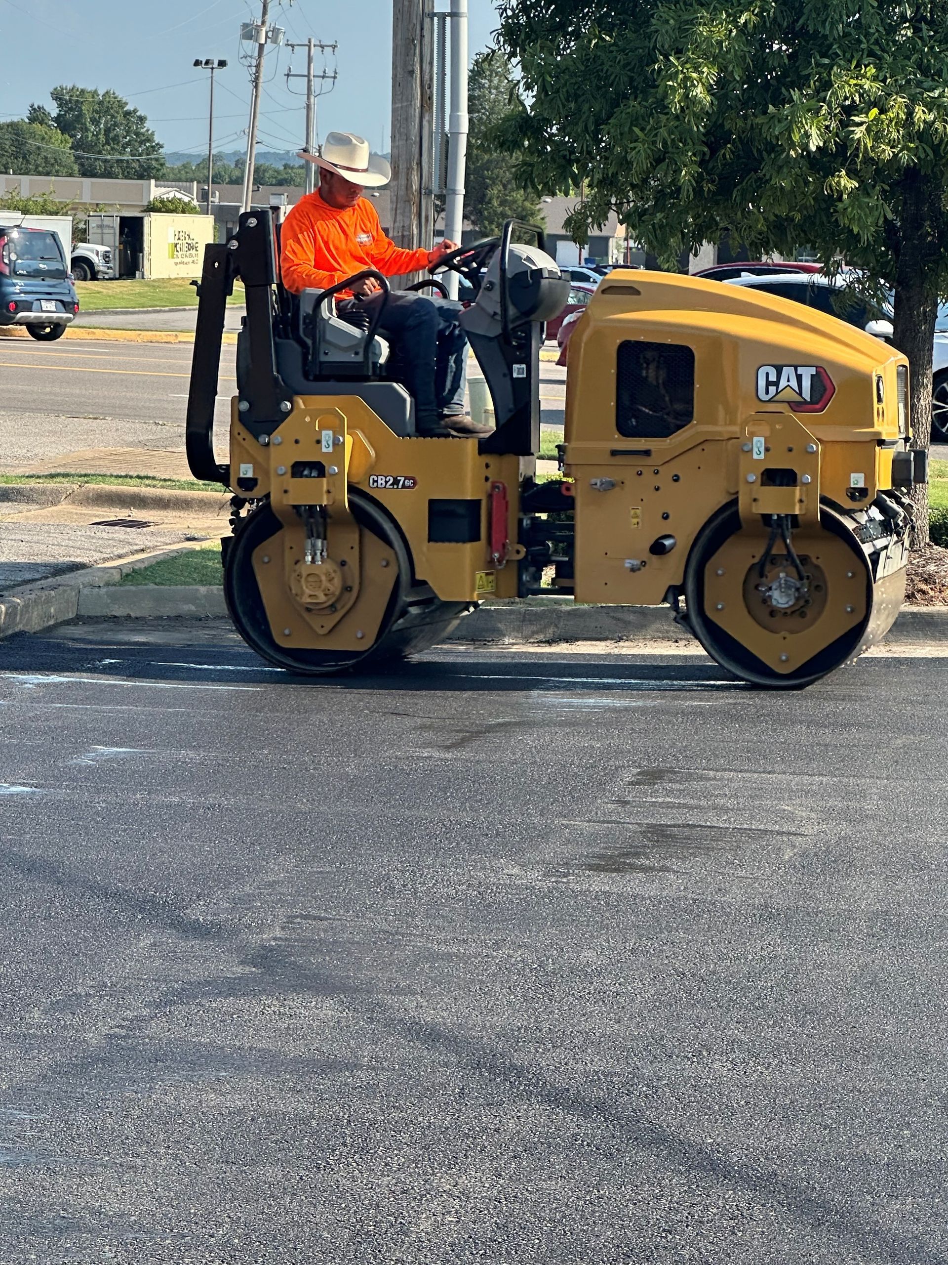 Person in orange shirt and cowboy hat operating a yellow CAT compactor, compacting asphalt.