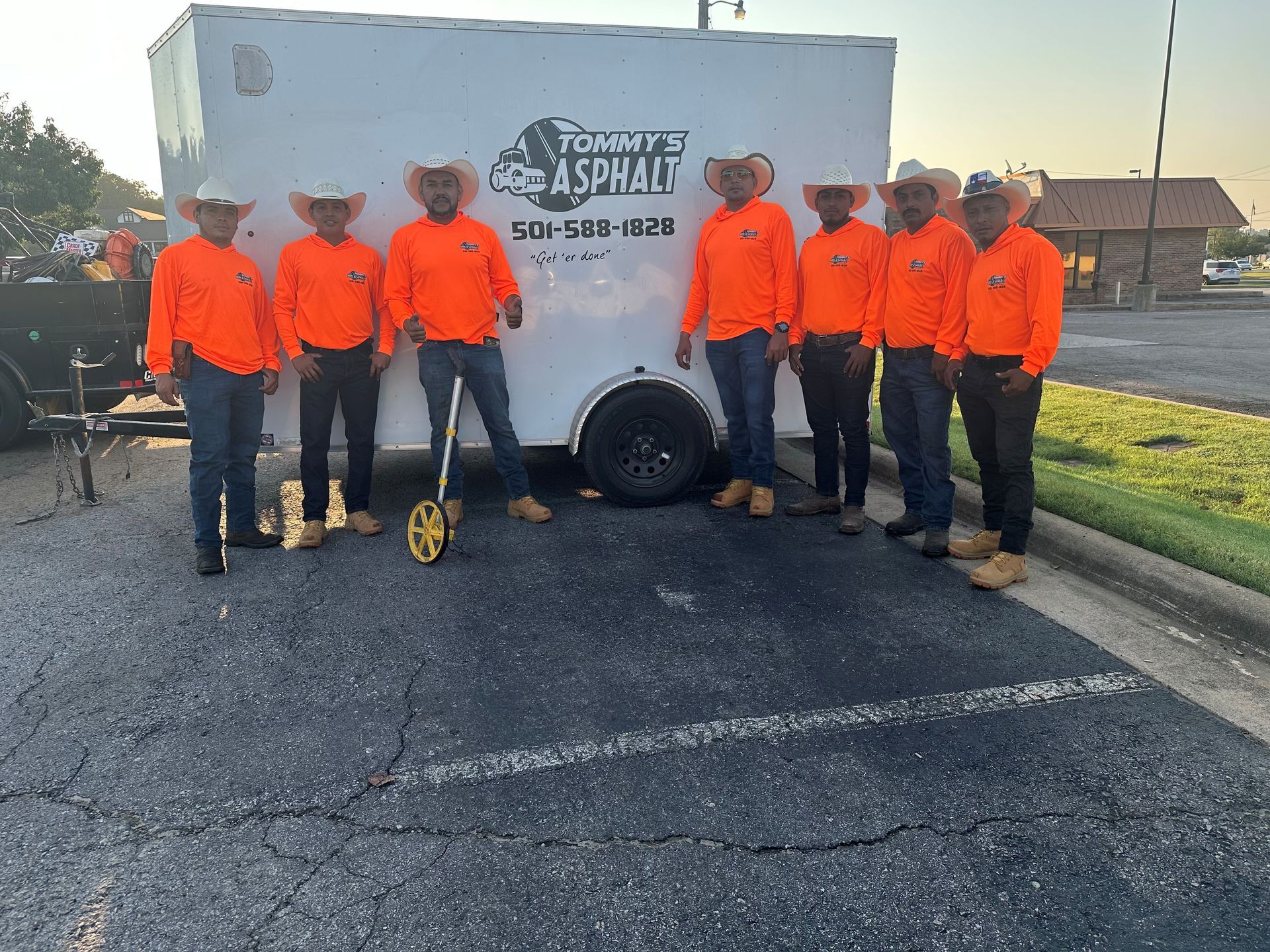 Group of asphalt workers in orange shirts and cowboy hats standing in front of trailer.