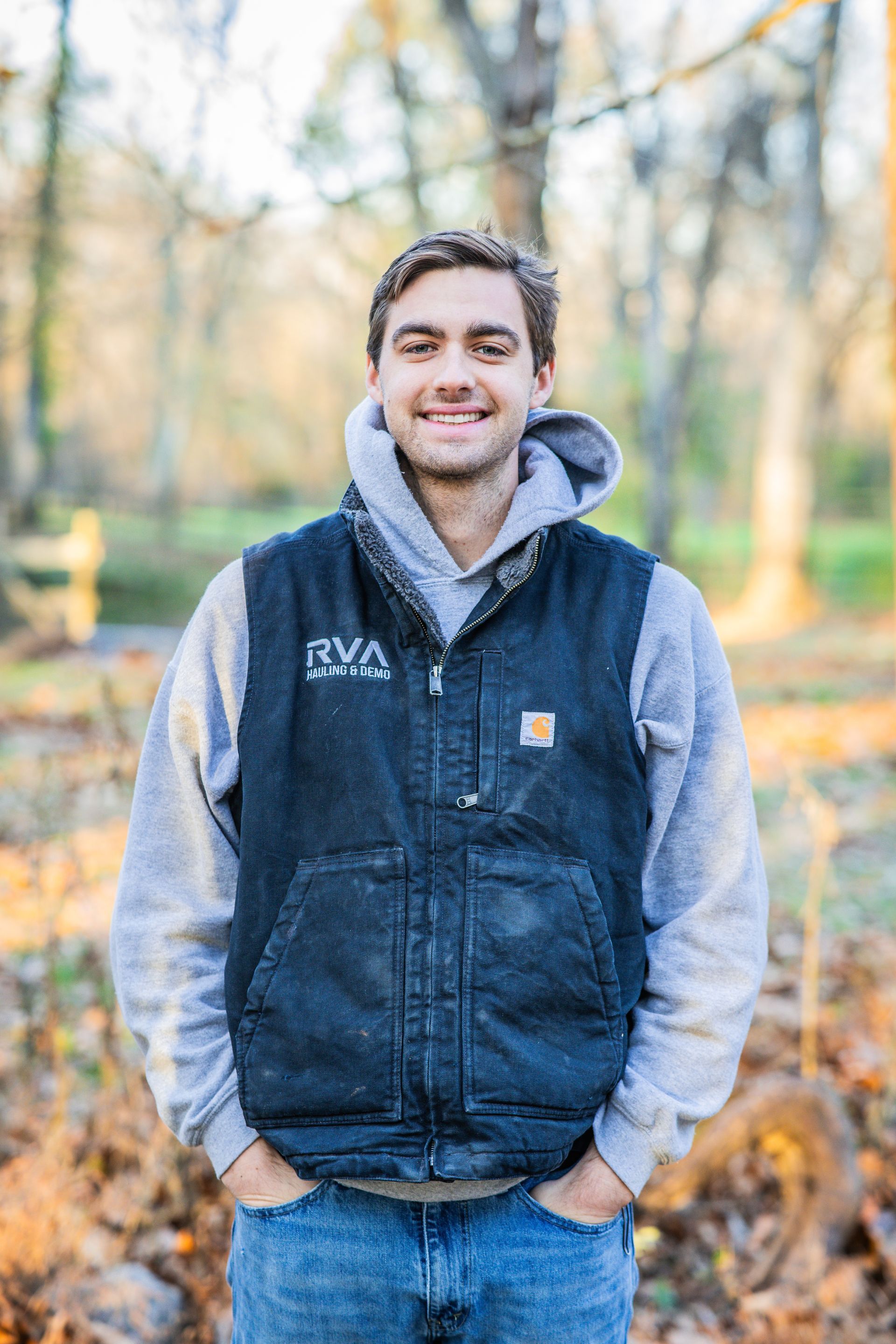 Man in grey hoodie and black vest smiles outdoors with hands in pockets.