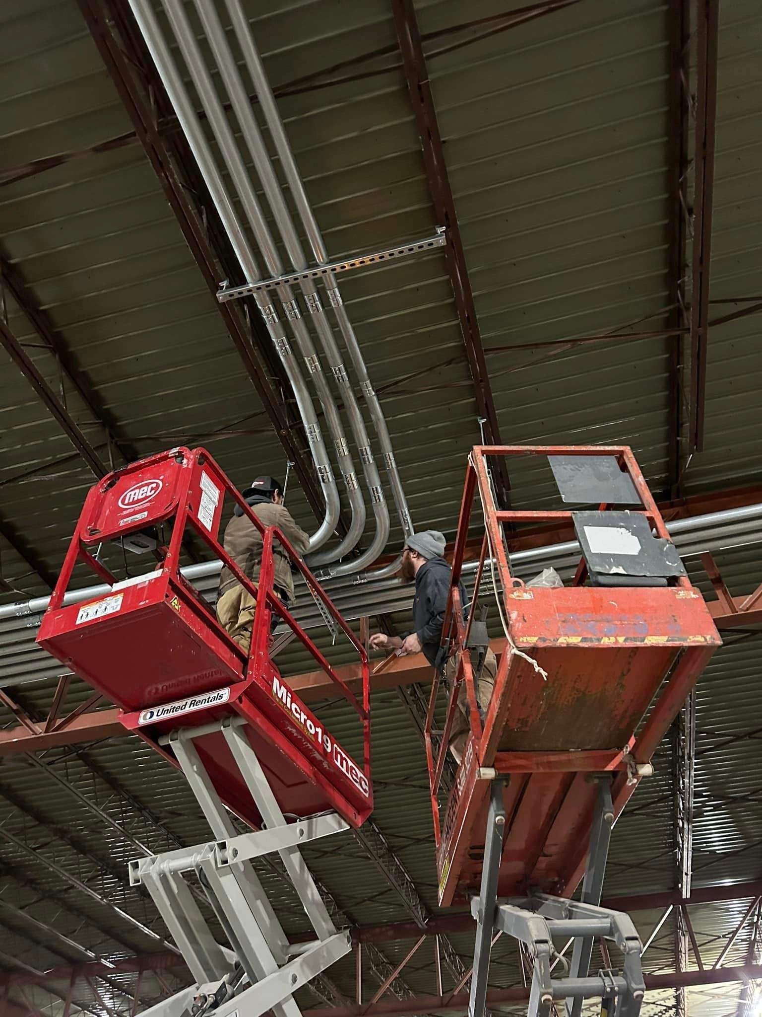 Two workers on a scissor lift are installing electrical wiring in the ceiling of an industrial facility