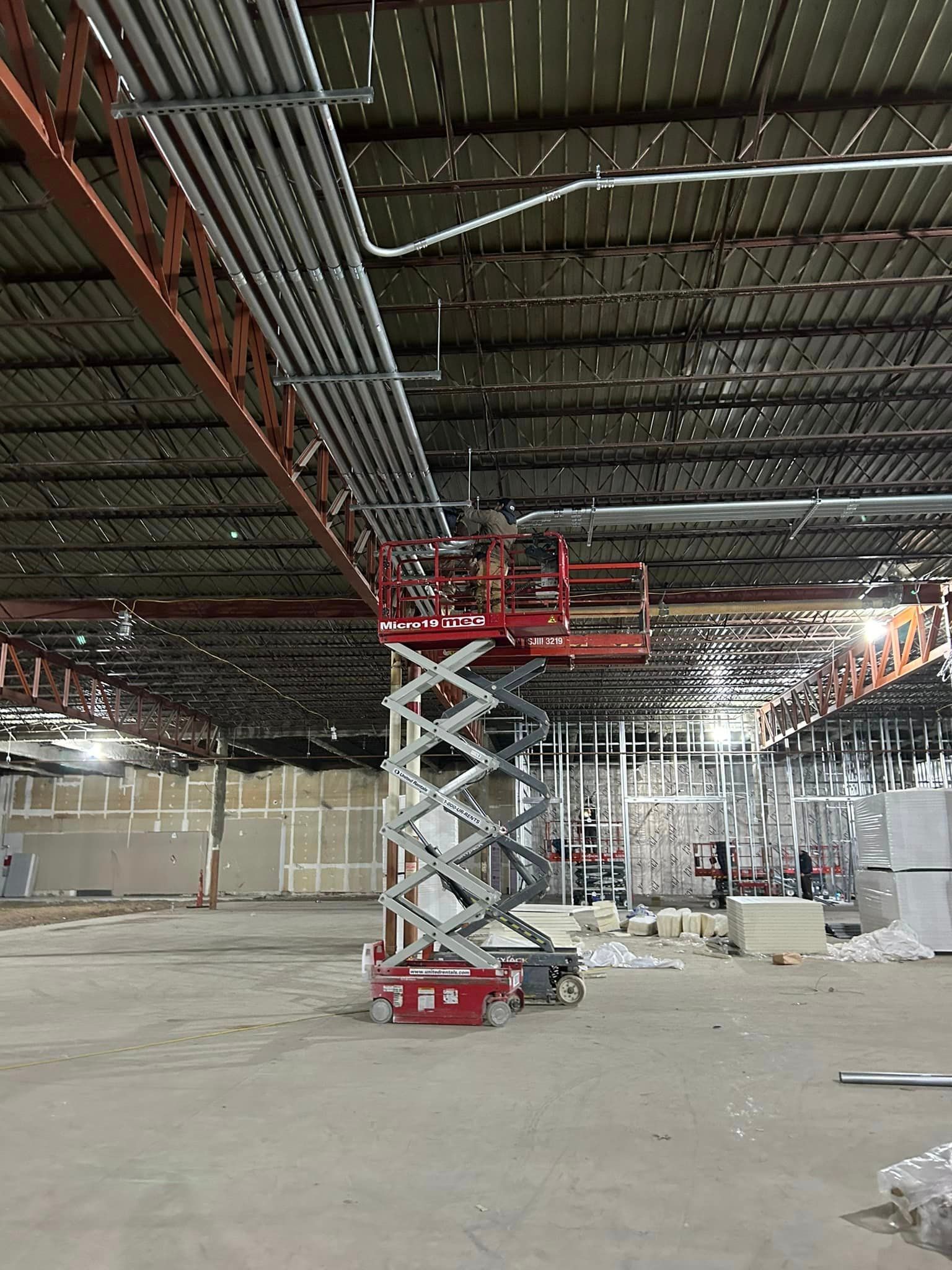 Two workers on a scissor lift are installing electrical wiring in the ceiling of an industrial unit