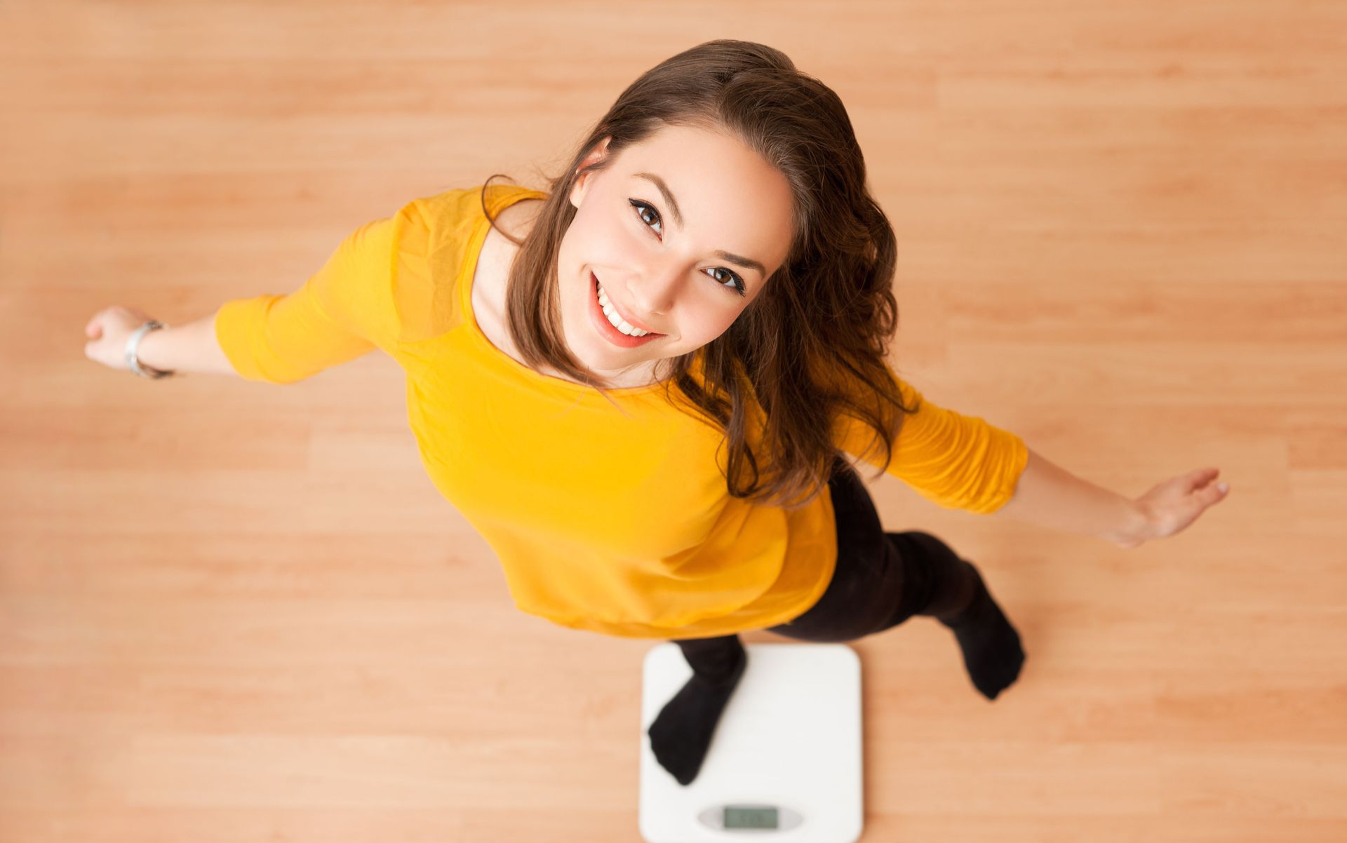 Woman smiles while standing on a scale; arms outspread. Light wood floor, wearing yellow shirt and black leggings.