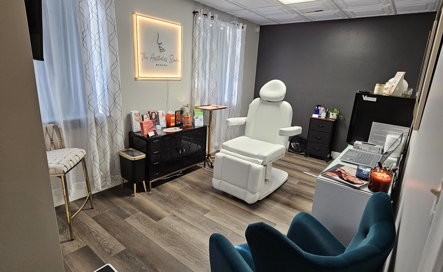 Aesthetic treatment room with chair, products, and waiting area. Gray and teal accents.