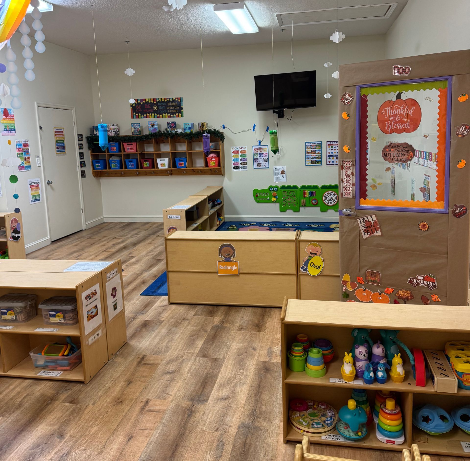 Interior of a child's classroom with wooden floors, shelves, a TV, and decorations.