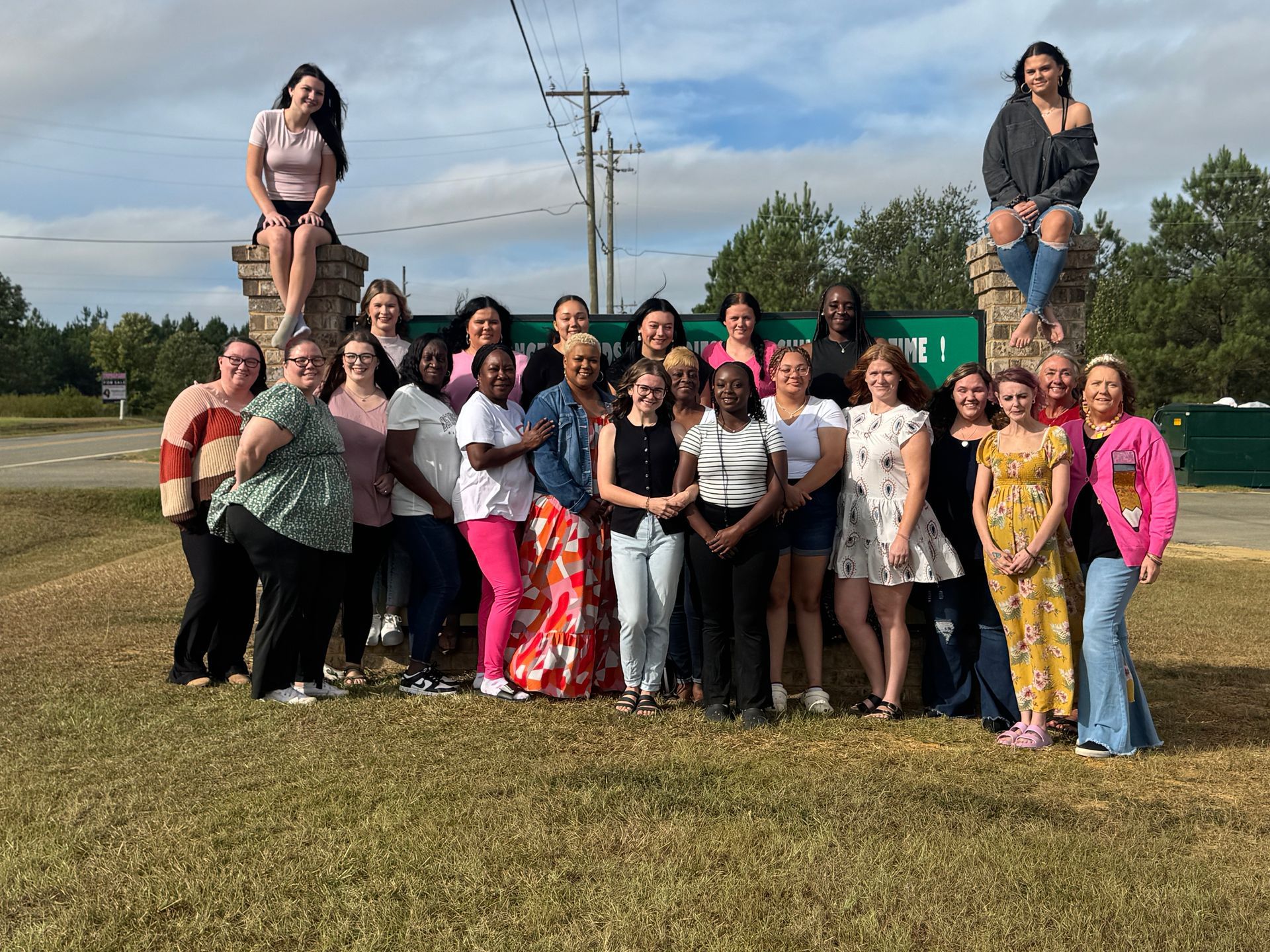 Group of women posing together near an entrance sign, some sitting on pillars..