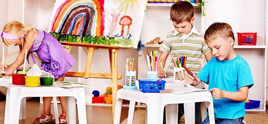 Children painting at tables in a brightly decorated art room; one painting on a large easel.