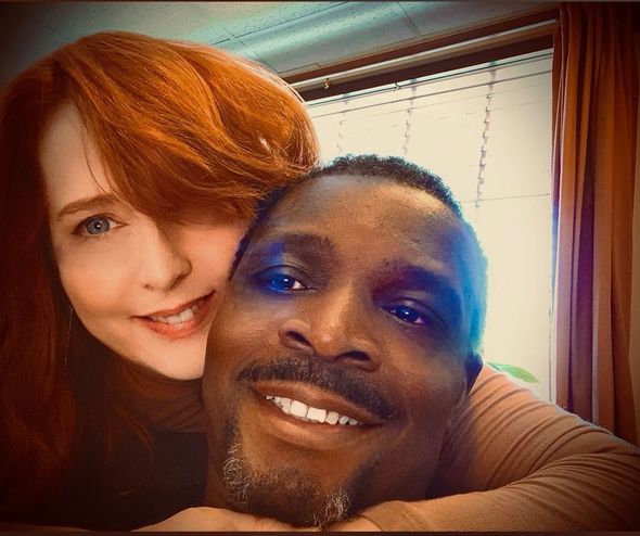A woman with red hair embraces a Black man, both smiling. Sunlight streams through a window.