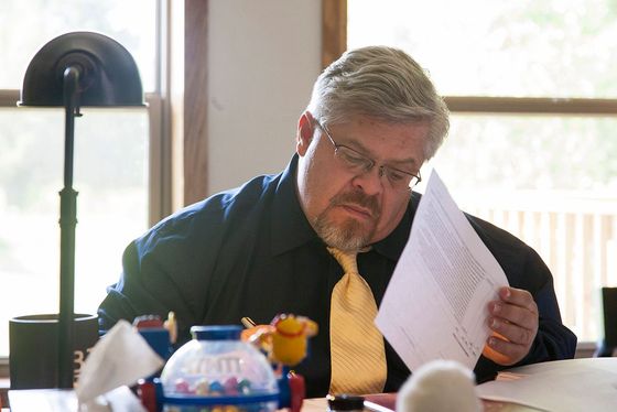 Man in glasses, yellow tie, reading papers at a desk near a window.